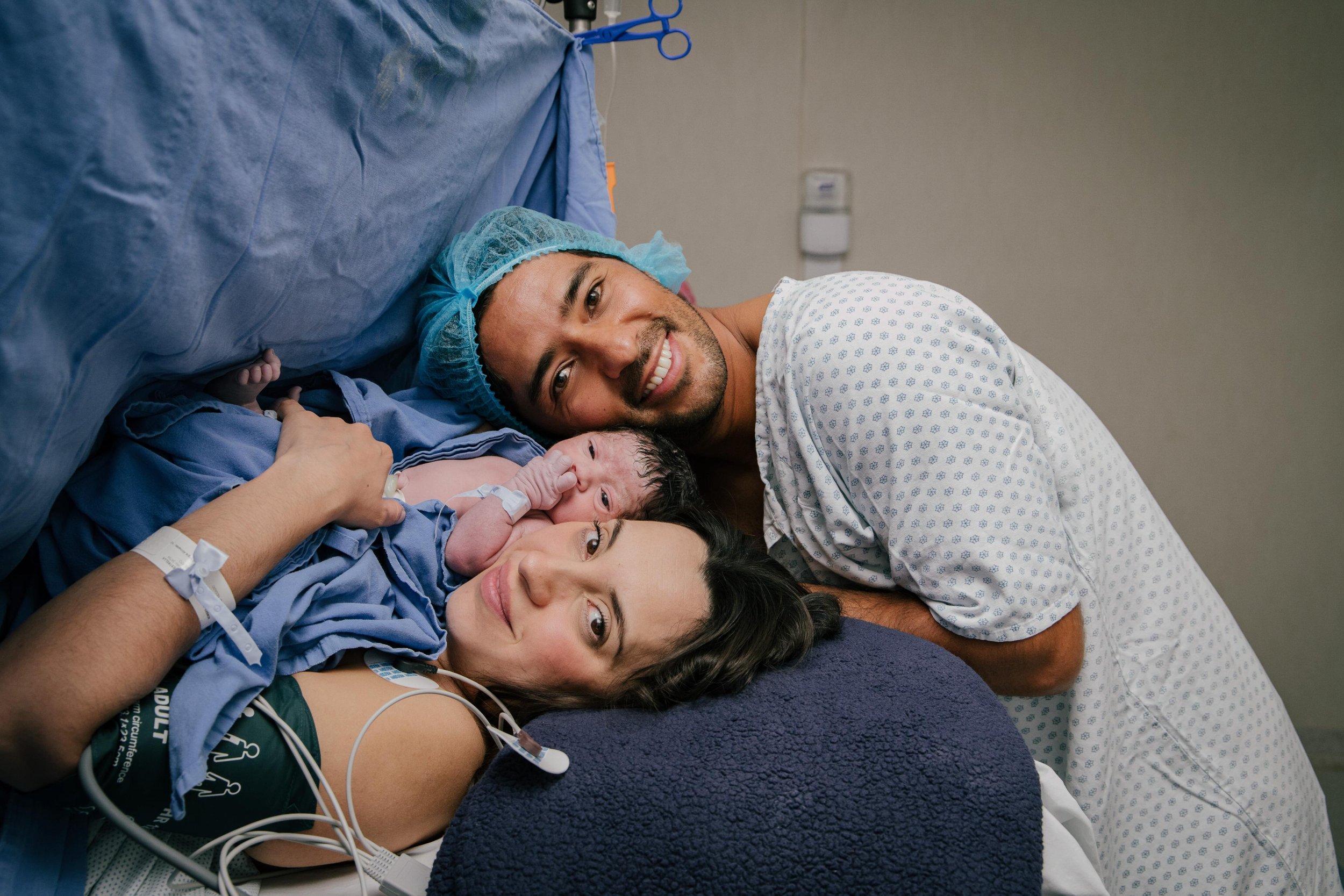Una pareja en un hospital con su recién nacido, todos sonrientes y rodeados de ambiente hospitalario. documentando su parto con fotografía de nacimiento.