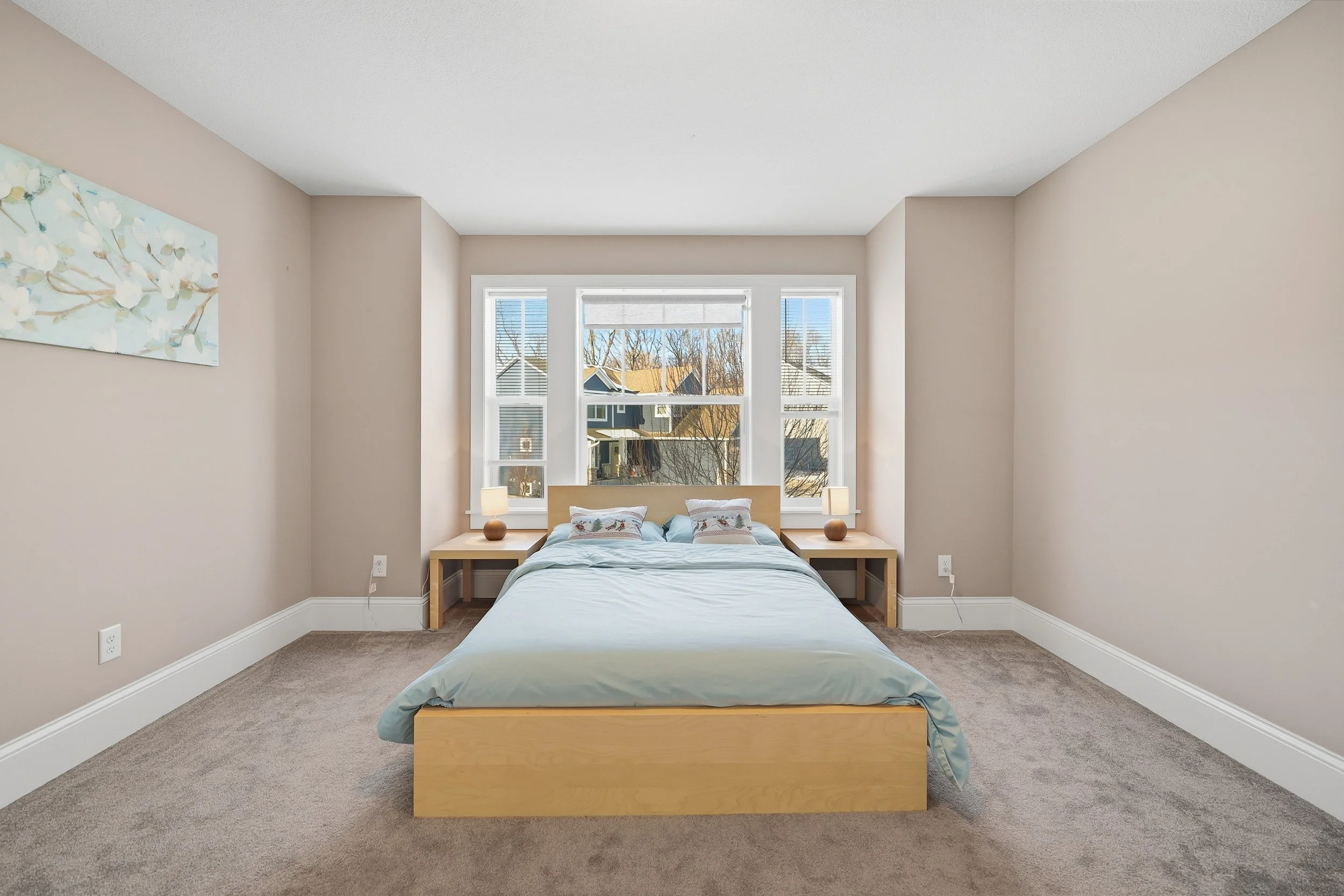 A bedroom with a large window, light-colored walls, a wooden bed with light blue bedding, and two nightstands with lamps.