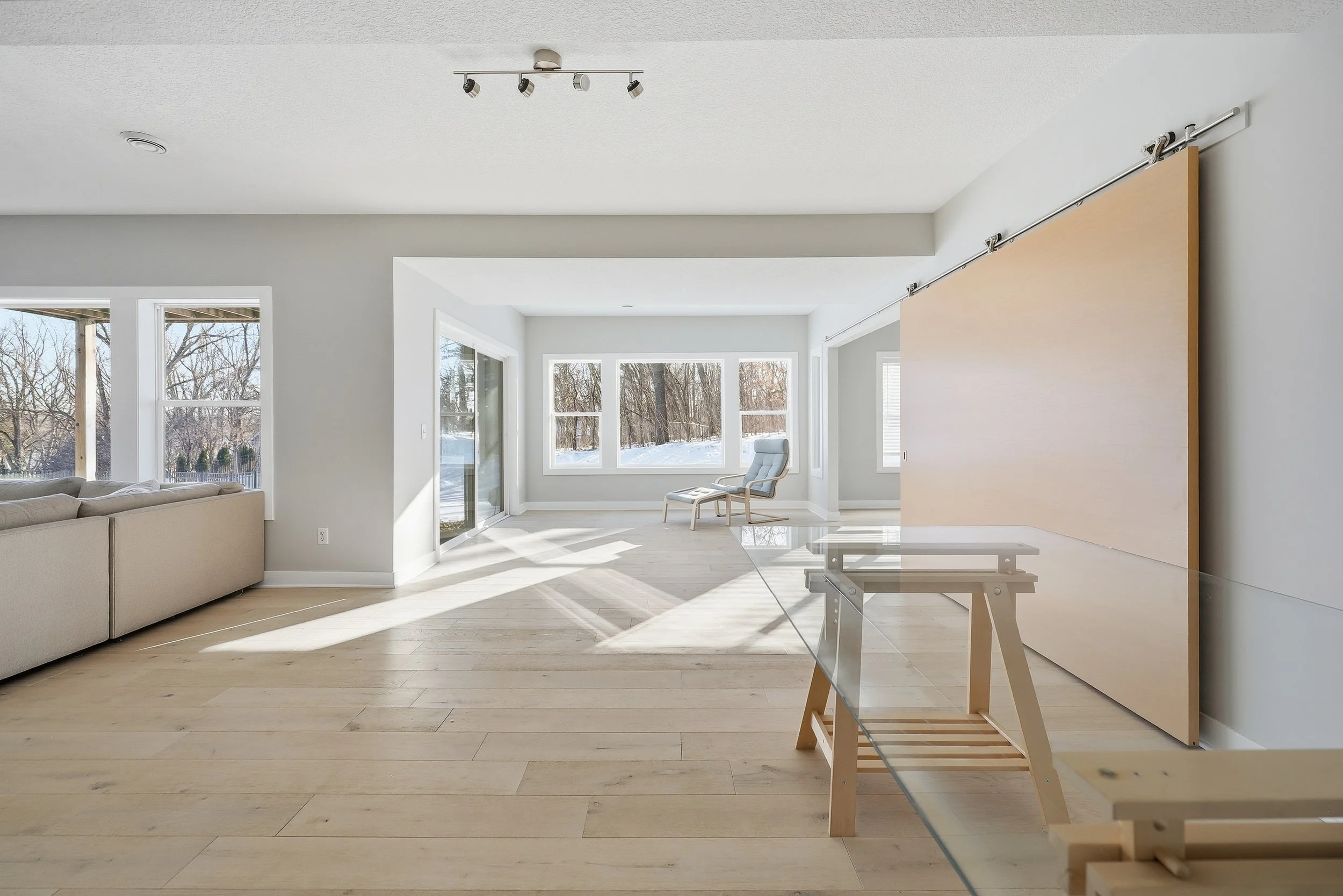 Empty living room with large windows, light-colored hardwood floors, a beige sofa, a lounge chair, and a sliding glass door leading outside.