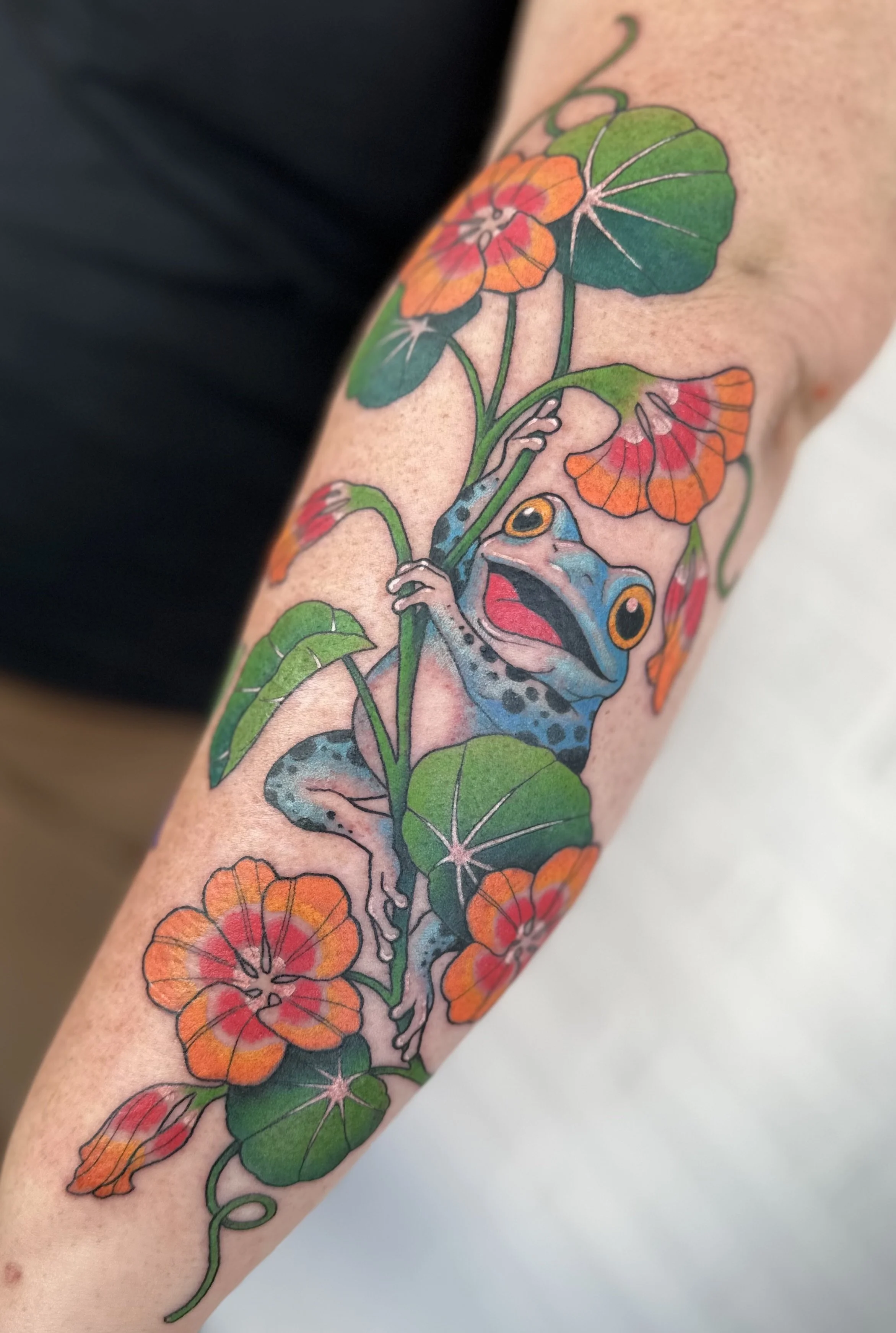 Färgglad tatuering av en groda som hänger i en växt med gröna blad och orange-röda blommor. Tatuering Visby