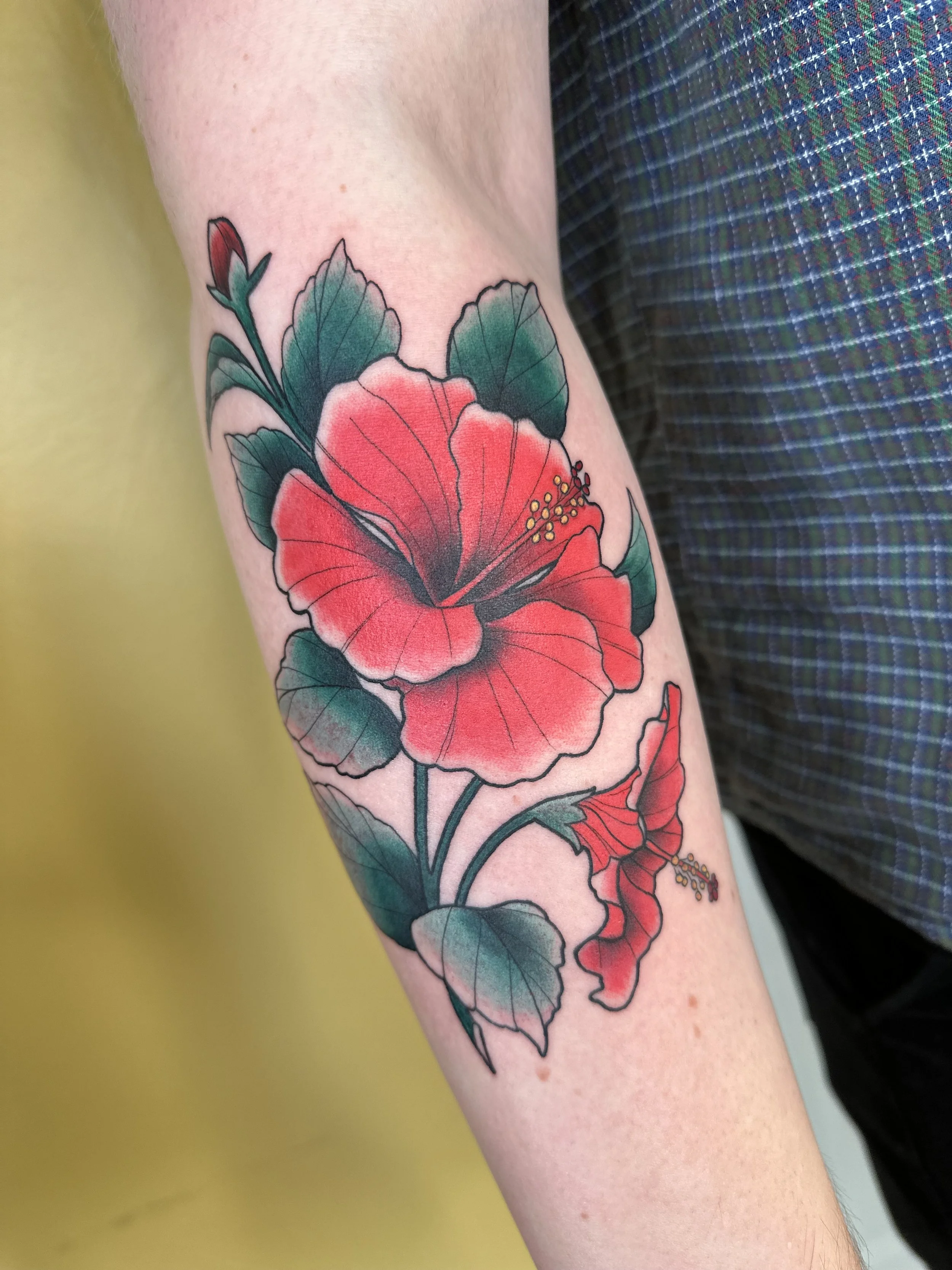 Färgstark tatuering av ett rött hibiskusblomma med gröna blad på en persons arm. Tatuering Visby