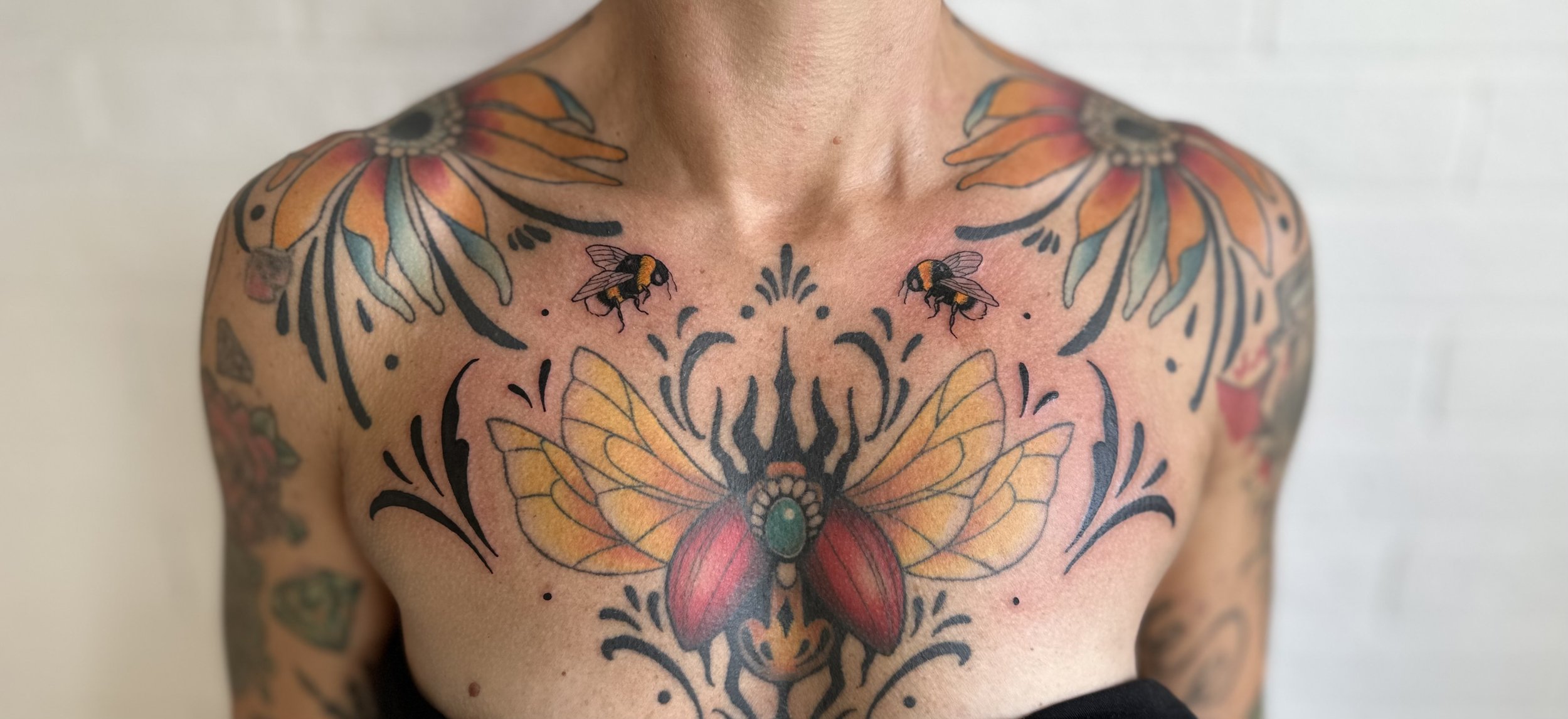 Färgglad tatuering på bröstet och axeln med en stor fjäril med gulaktiga vingar och röda kropp, två bin nära fjärilen, och blommor och dekorativa mönster Chestpiece skalbagge bi tatuering Visby Gotland 