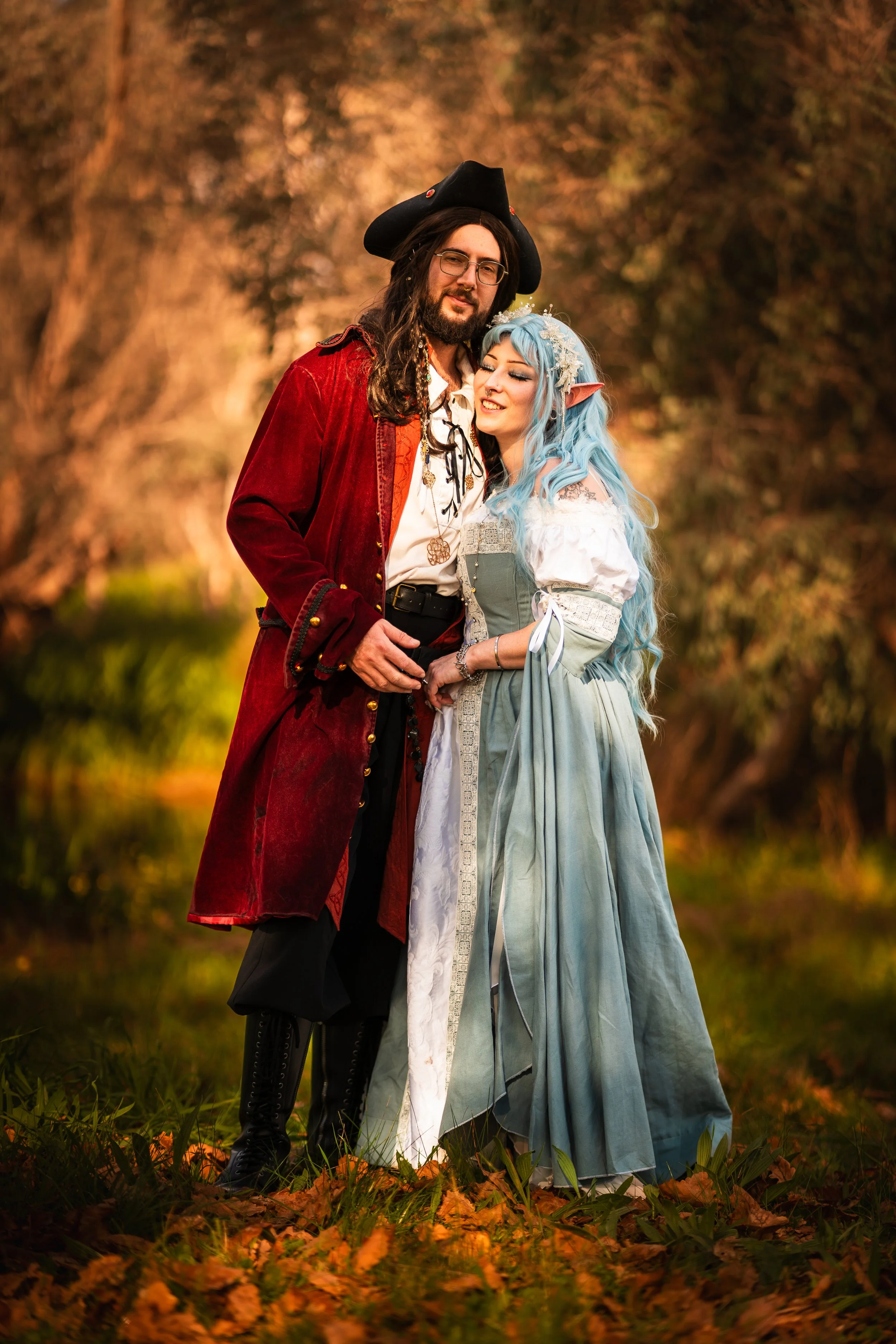 A man dressed as a pirate and a woman dressed as an elf stand close together outdoors during autumn, surrounded by fallen leaves and trees with golden foliage.