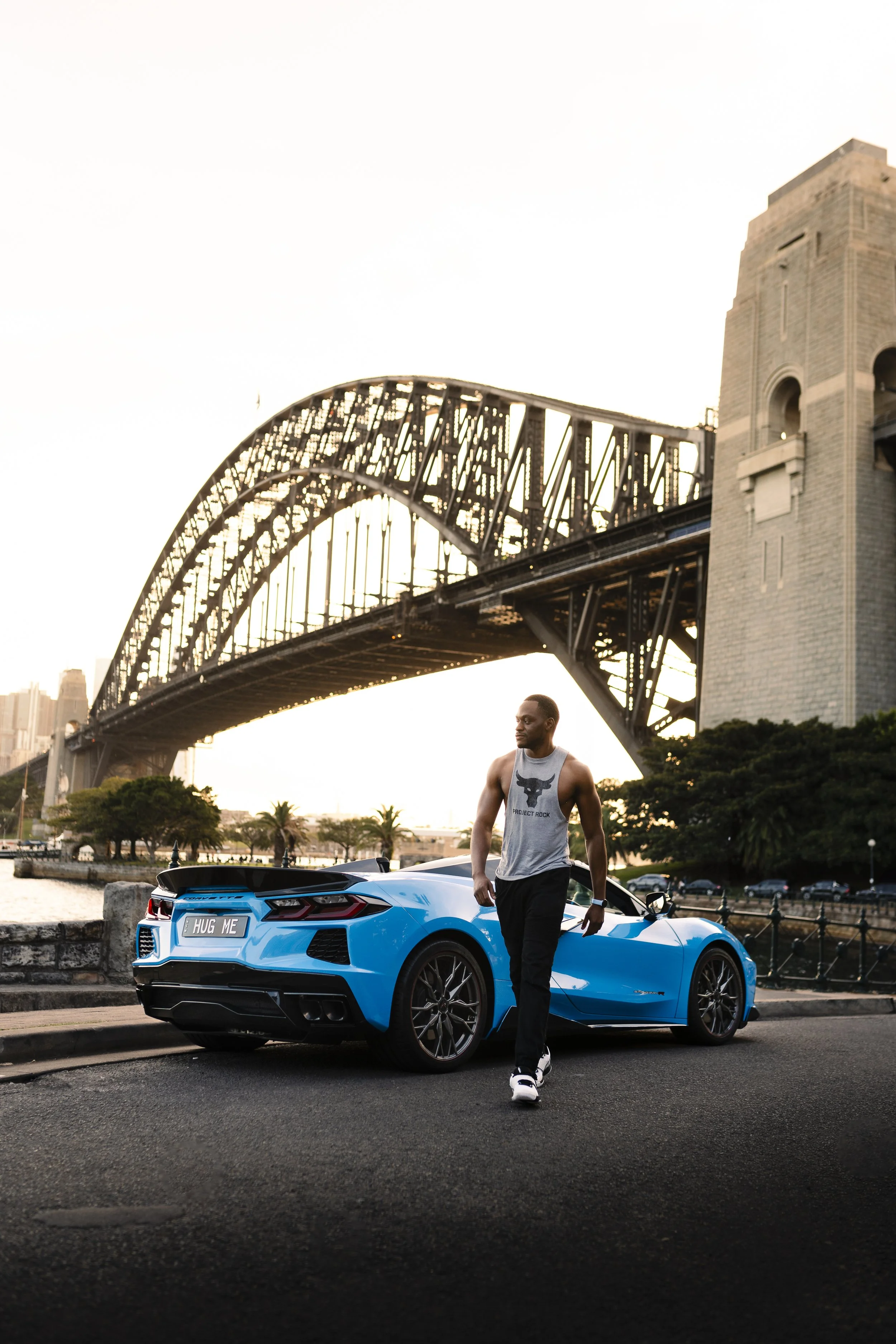 A man in a gray tank top and black pants walking beside a blue sports car with a custom license plate 'HUG ME' under the Sydney Harbour Bridge in Australia during sunset.