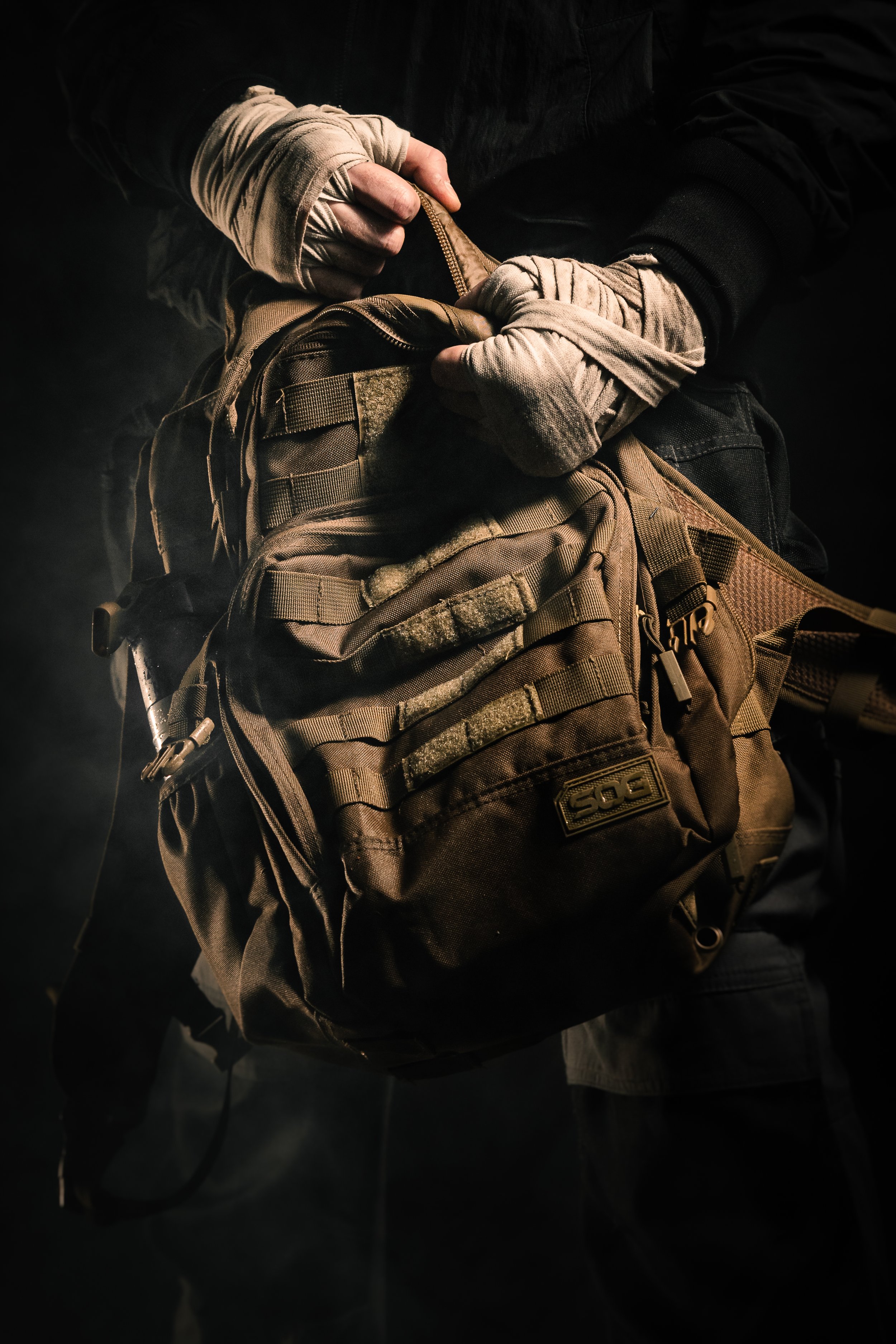 Person wearing tactical gloves holds a brown military-style backpack in a dark setting.