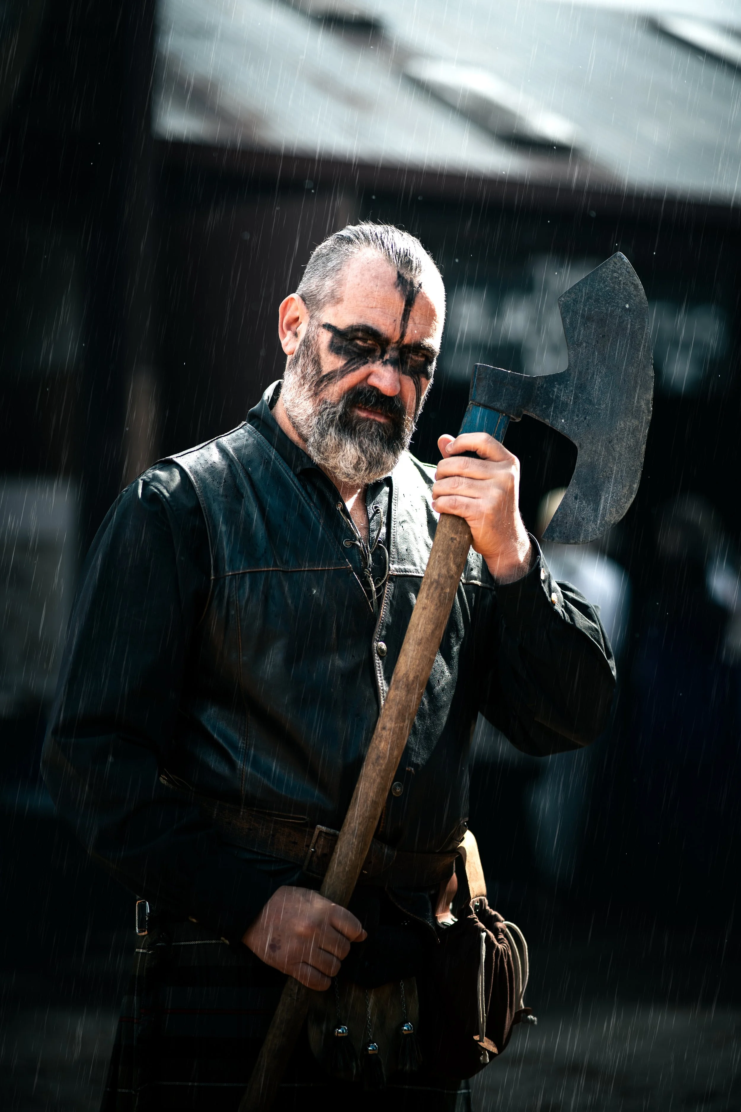 A man with black paint on face, wearing a leather jacket, holding an axe in the rain.