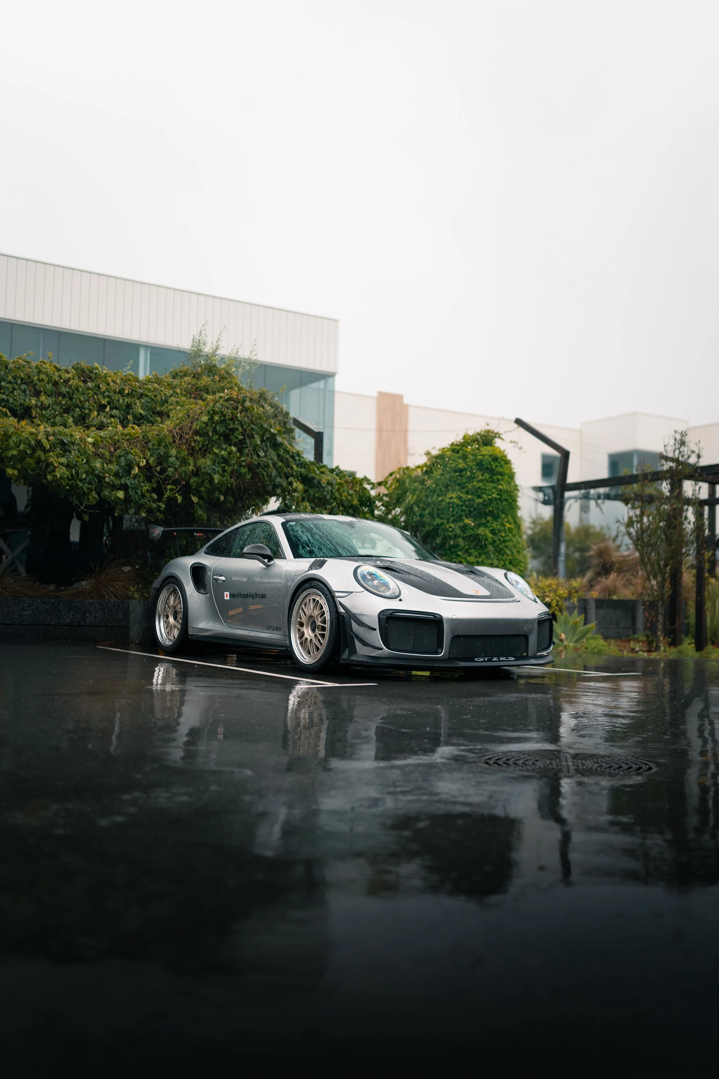 A silver Porsche 911 GT2 RS parked on a wet pavement, with greenery and modern buildings in the background on a rainy day.