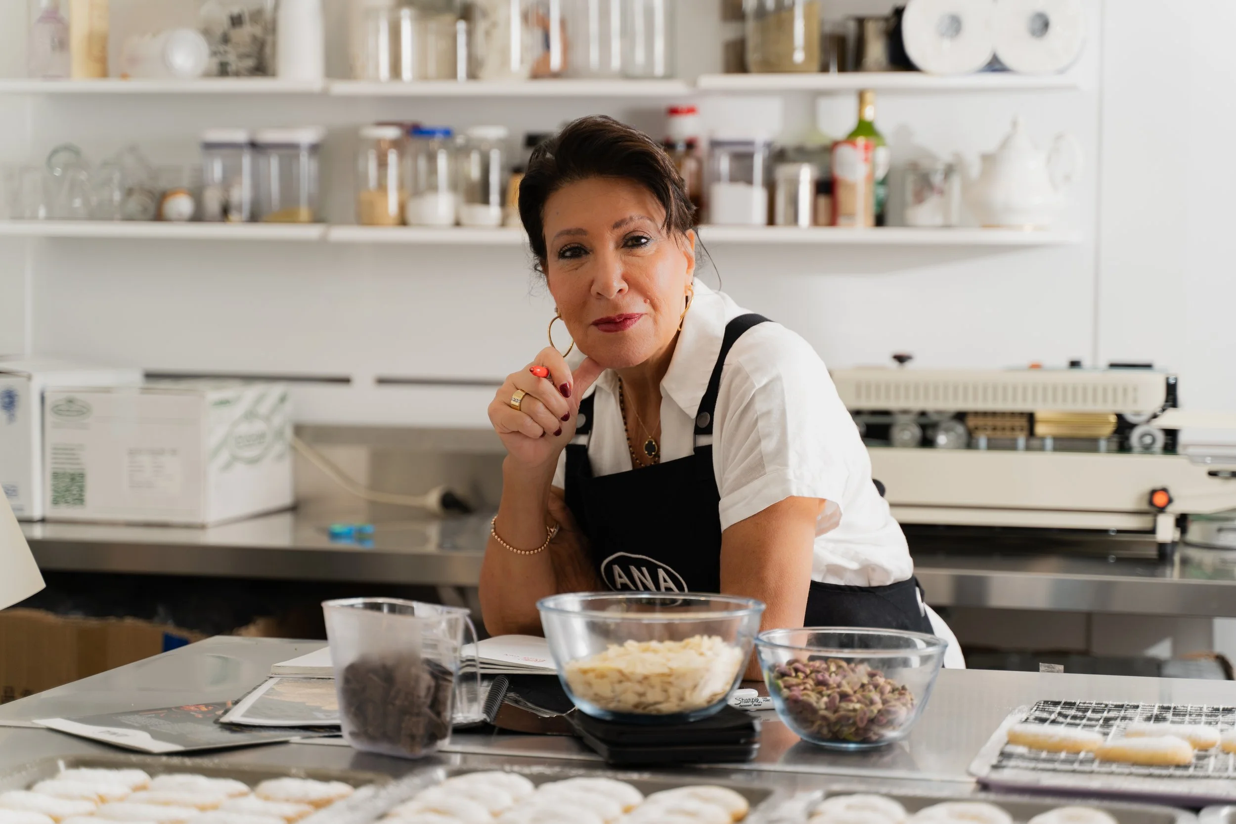 A woman with dark hair, wearing a white shirt and black apron, sitting at a kitchen counter with baking ingredients and cookies in front of her.