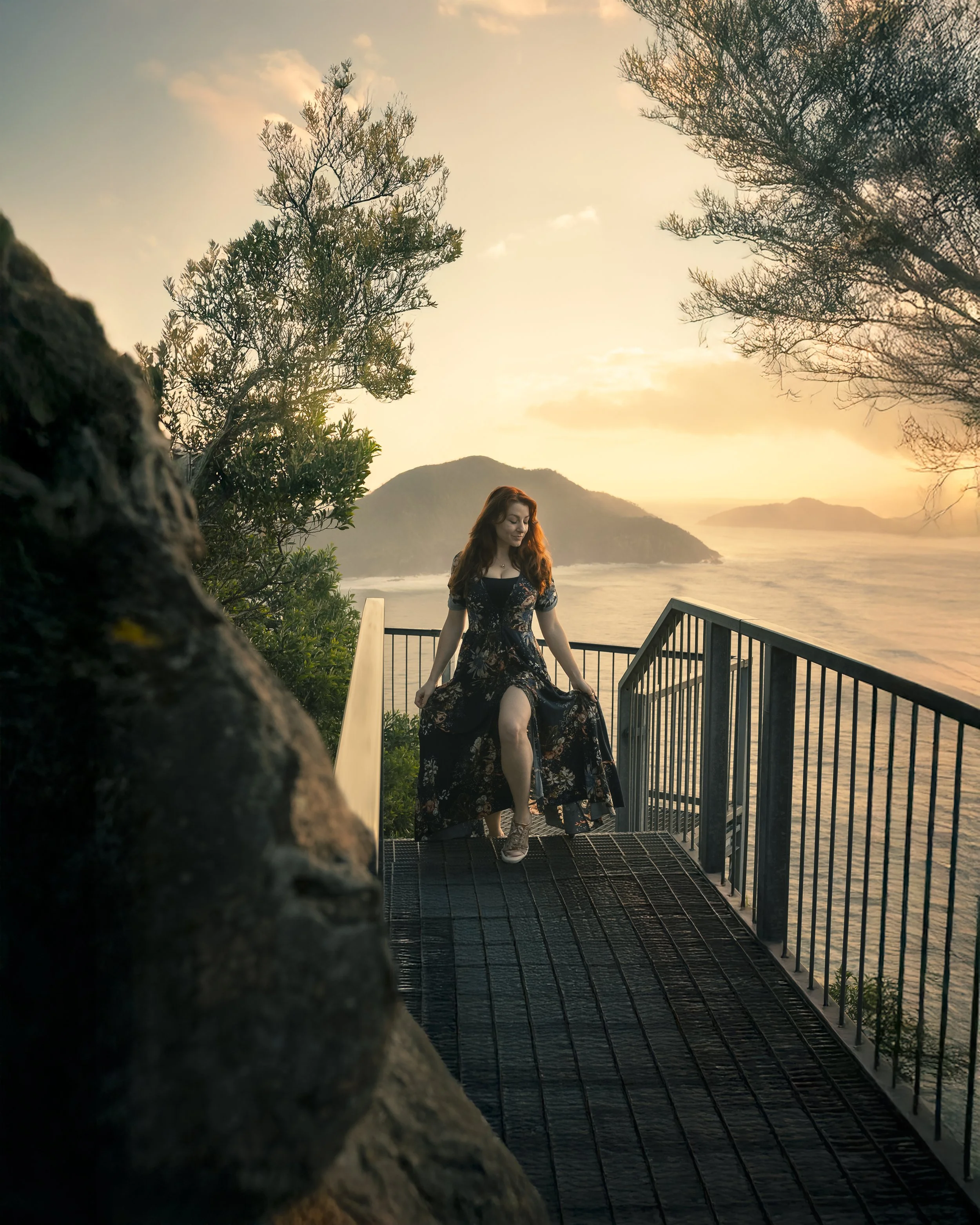 A woman in a long floral dress walking up a metal staircase towards a scenic view of the ocean at sunset, with islands in the background and trees around.