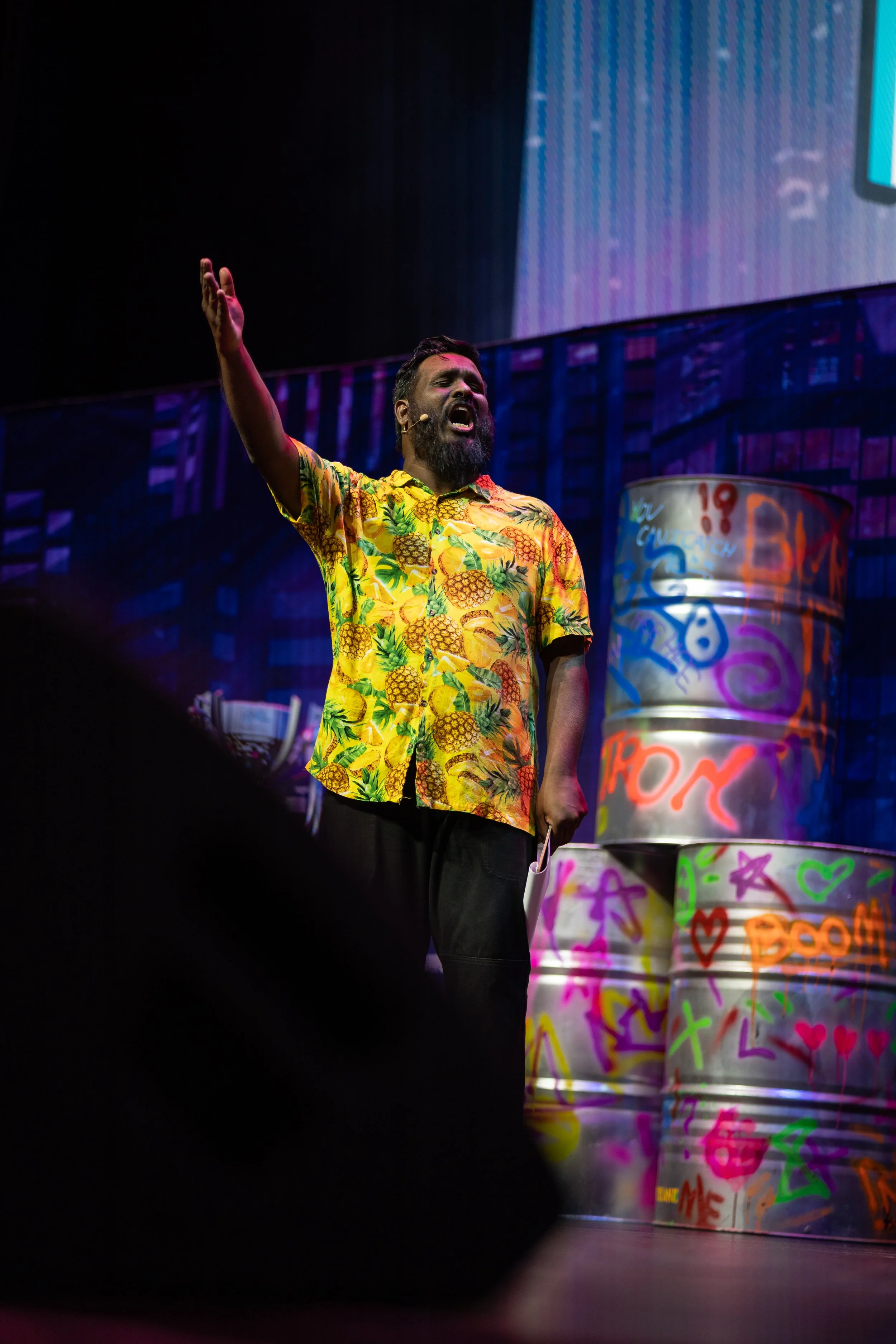 A man with a beard wearing a yellow Hawaiian shirt with pineapples is speaking on stage with his right arm raised. Behind him are four colorful, graffiti-covered drums.