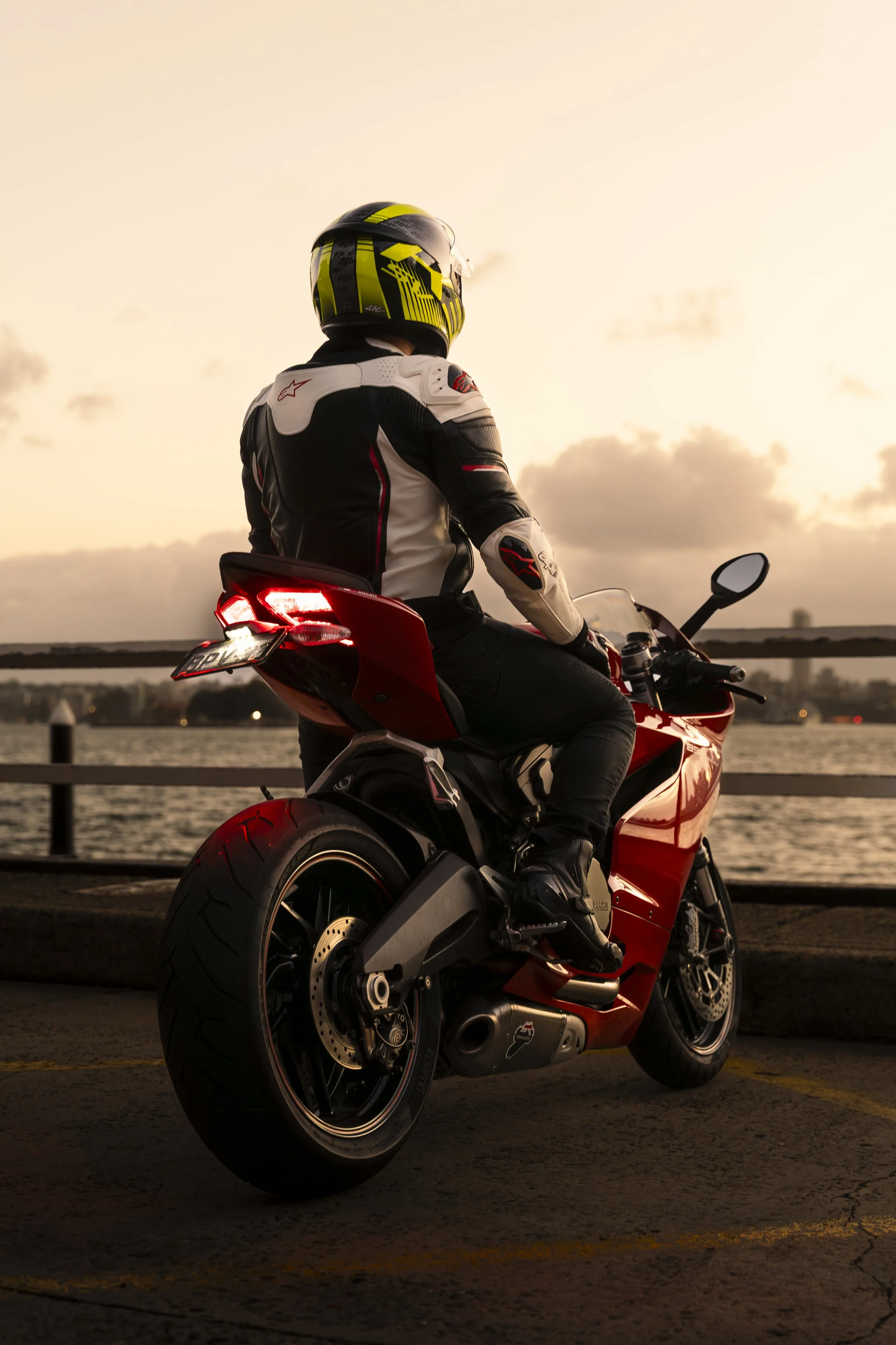 Motorcycle rider in racing gear sitting on red motorcycle by waterfront during sunset.