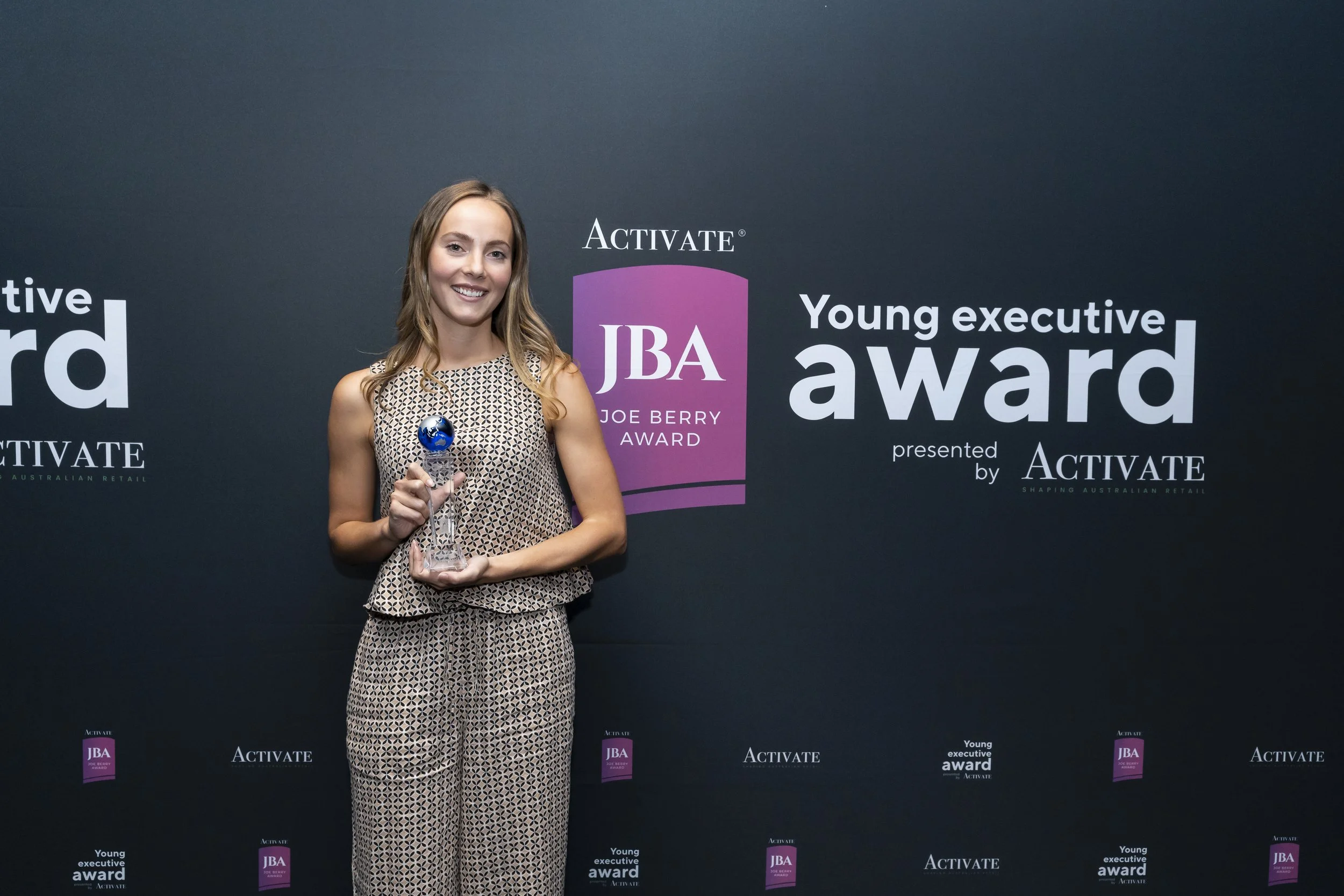 Young woman in patterned outfit holding an award while standing in front of a backdrop with logos for the Young Executive Award, the JBA Joe Berry Award, and Activate.