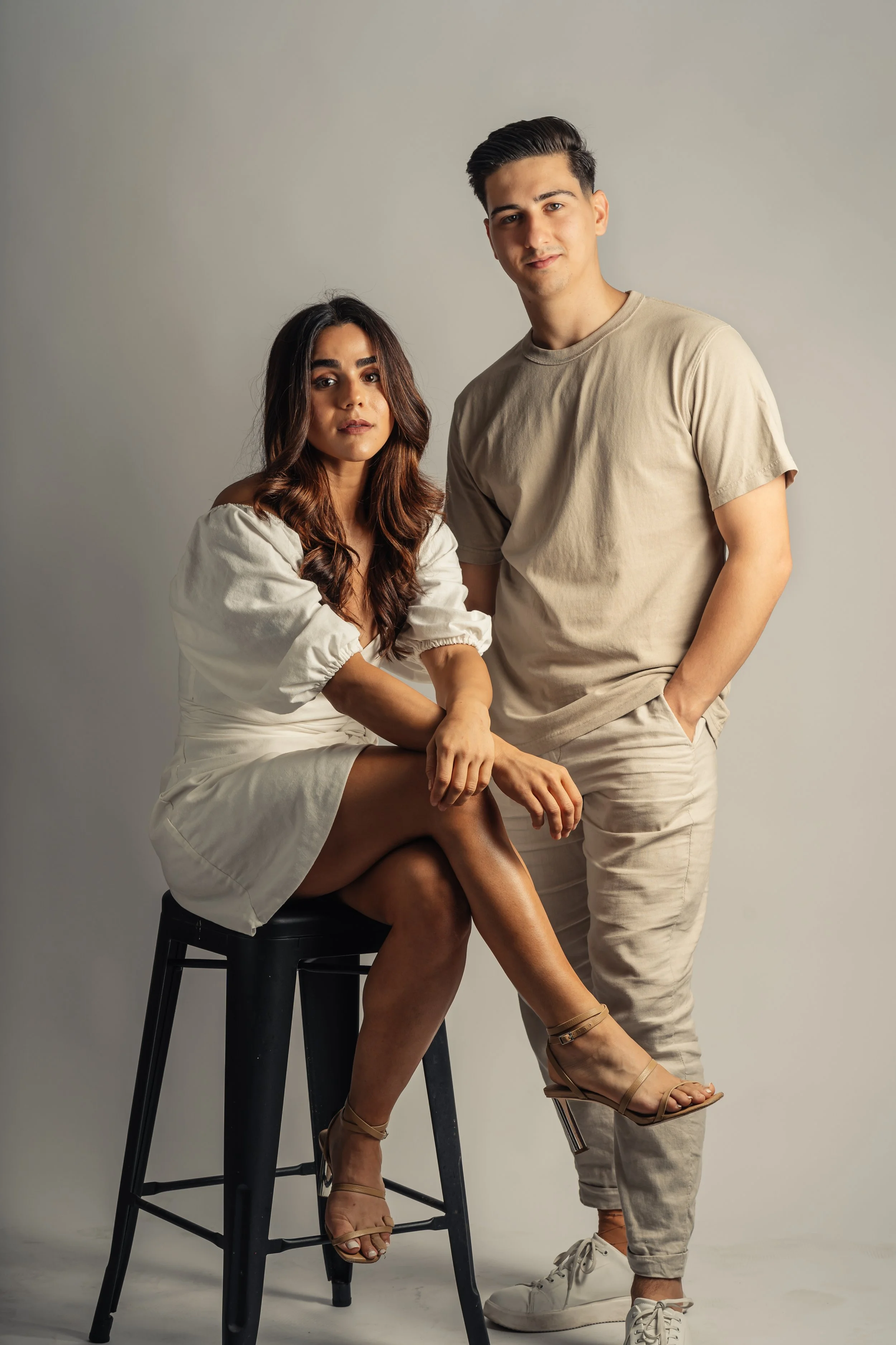 A man and a woman posing together against a plain neutral background. The woman is seated on a black stool with her legs crossed, wearing a white dress and beige high heels. The man is standing beside her with one hand in his pocket, dressed in a beige t-shirt, beige pants, and white sneakers.