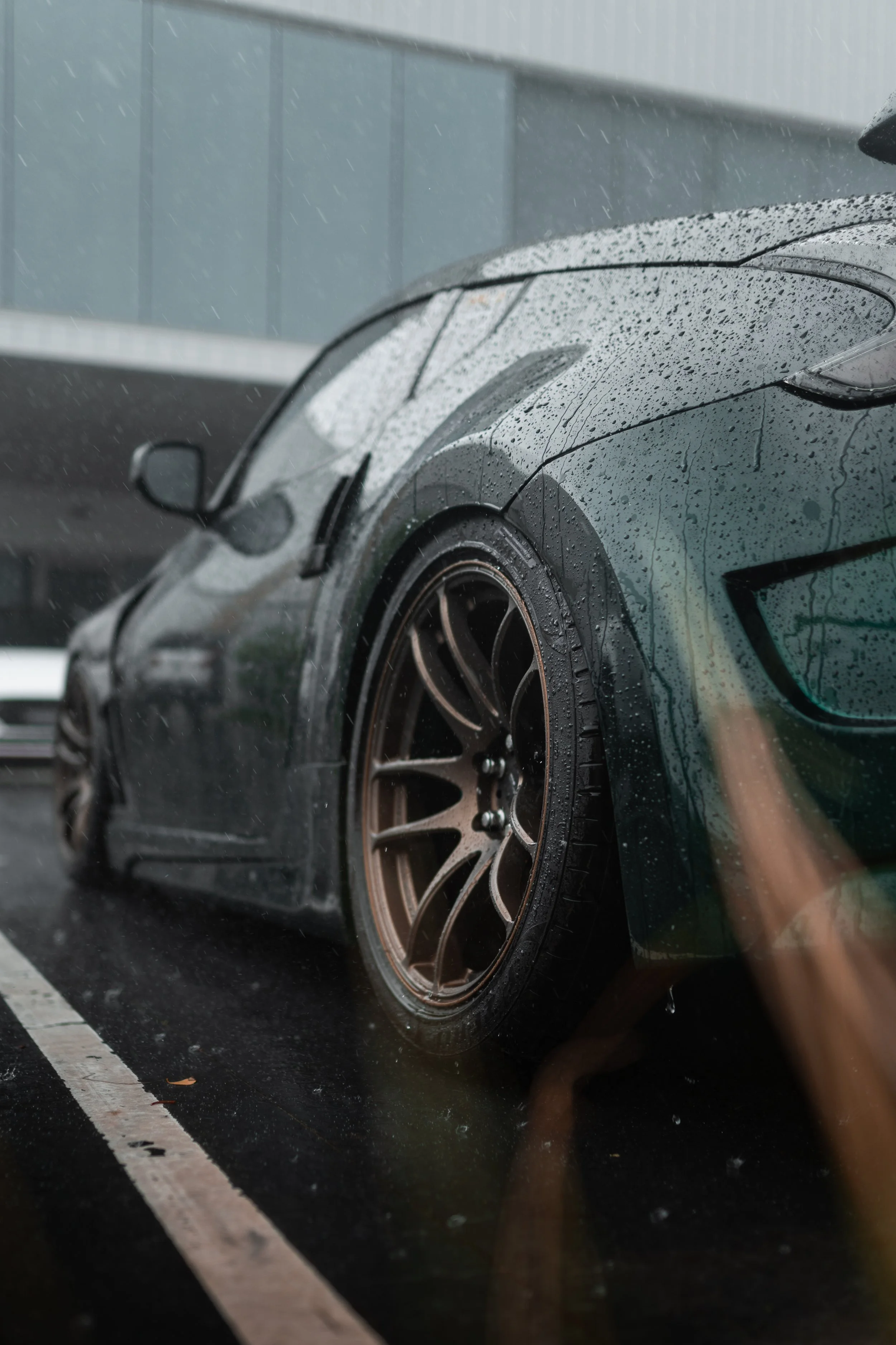 A dark-colored sports car with bronze wheels parked in the rain, with water droplets on the car's surface and wet pavement.