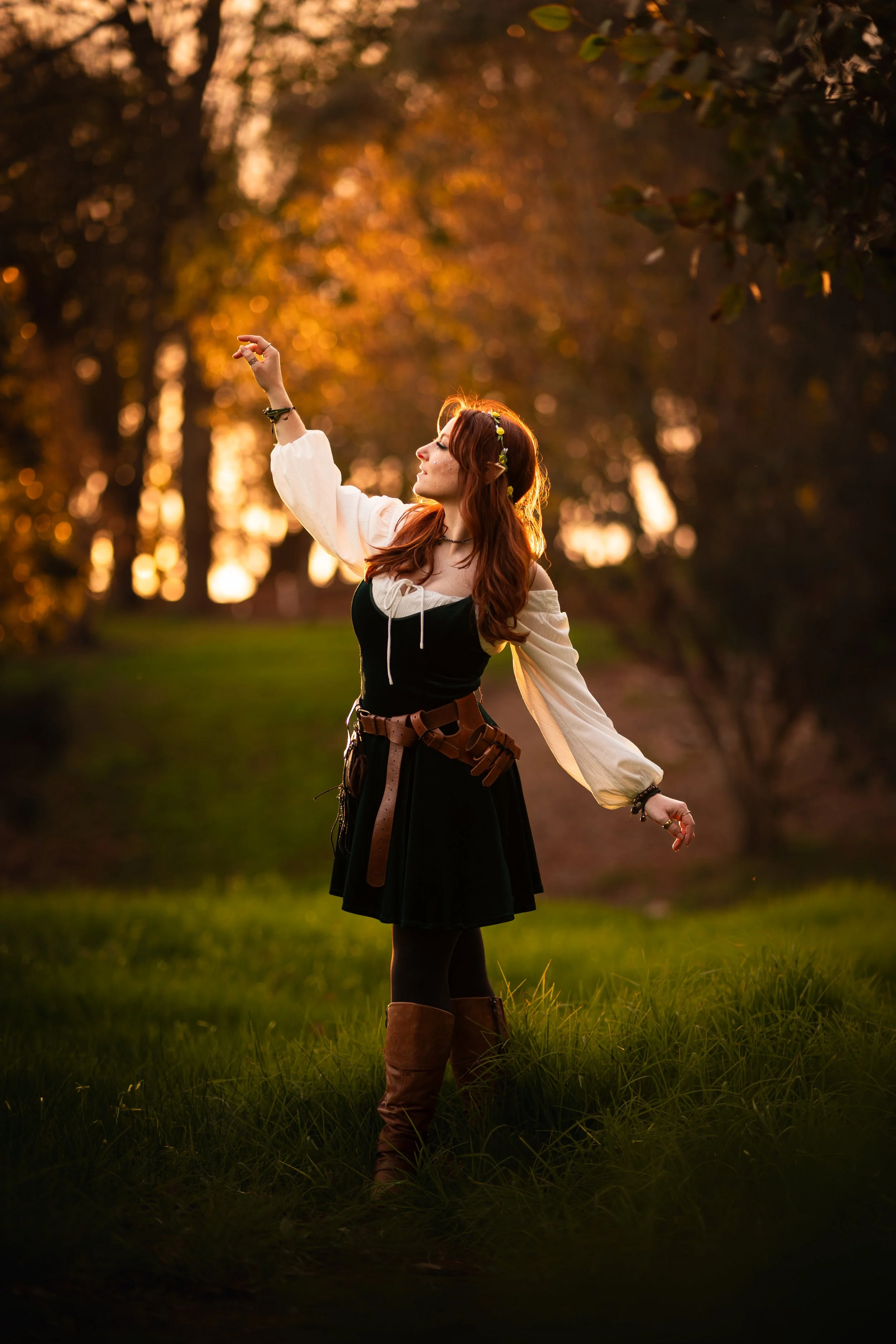 A woman with red hair, wearing a black dress with white sleeves, a brown belt, and tall brown boots, dancing or posing outdoors at sunset, surrounded by greenery and trees.