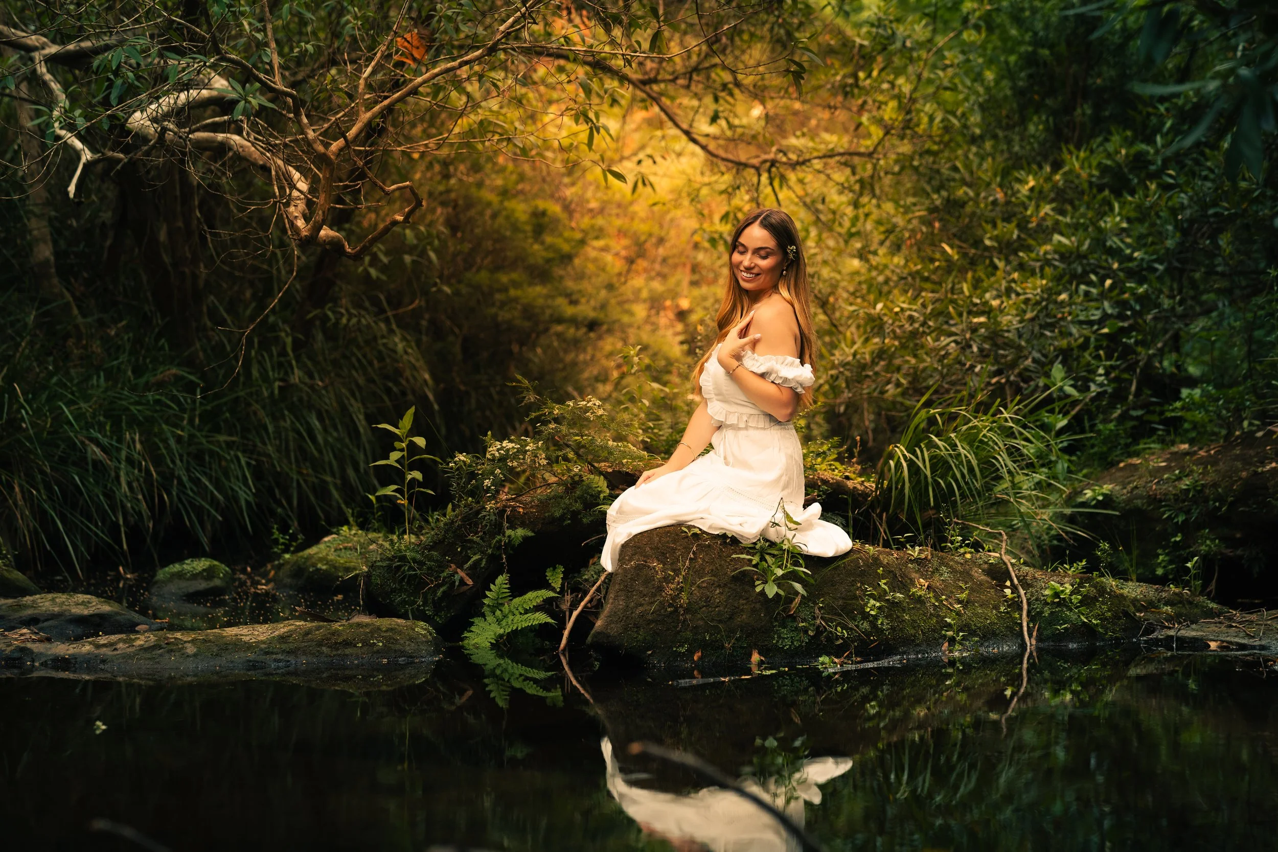 A young woman in a white dress sitting on a mossy rock beside a small stream in a lush green forest.