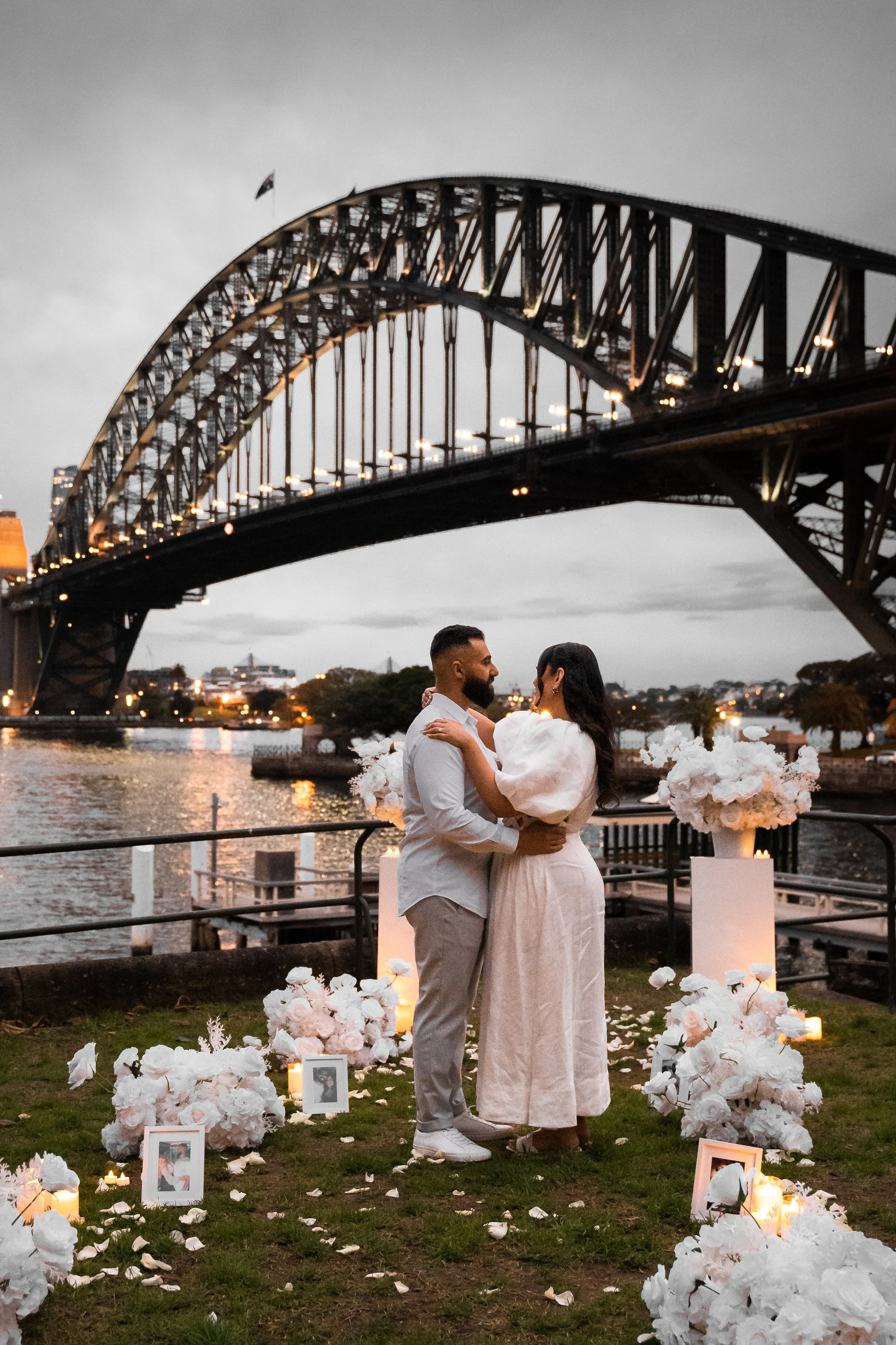 A couple in wedding attire stands close together during their outdoor wedding ceremony near a river, under the Sydney Harbour Bridge at dusk, surrounded by white floral arrangements, candles, and photographs.