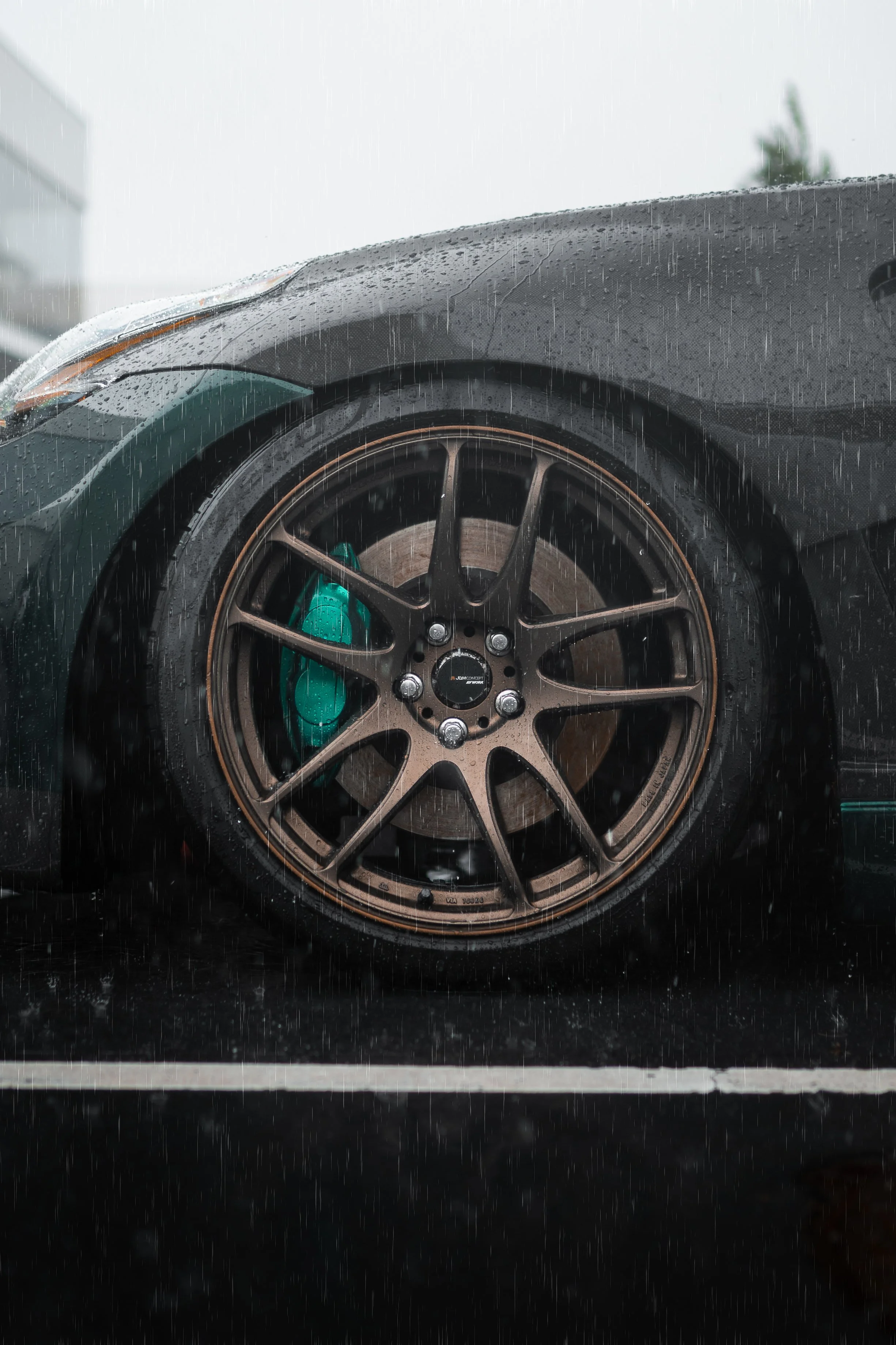 Close-up of a car wheel with a bronze multi-spoke rim and a green brake caliper, on a rainy day at a parking lot.
