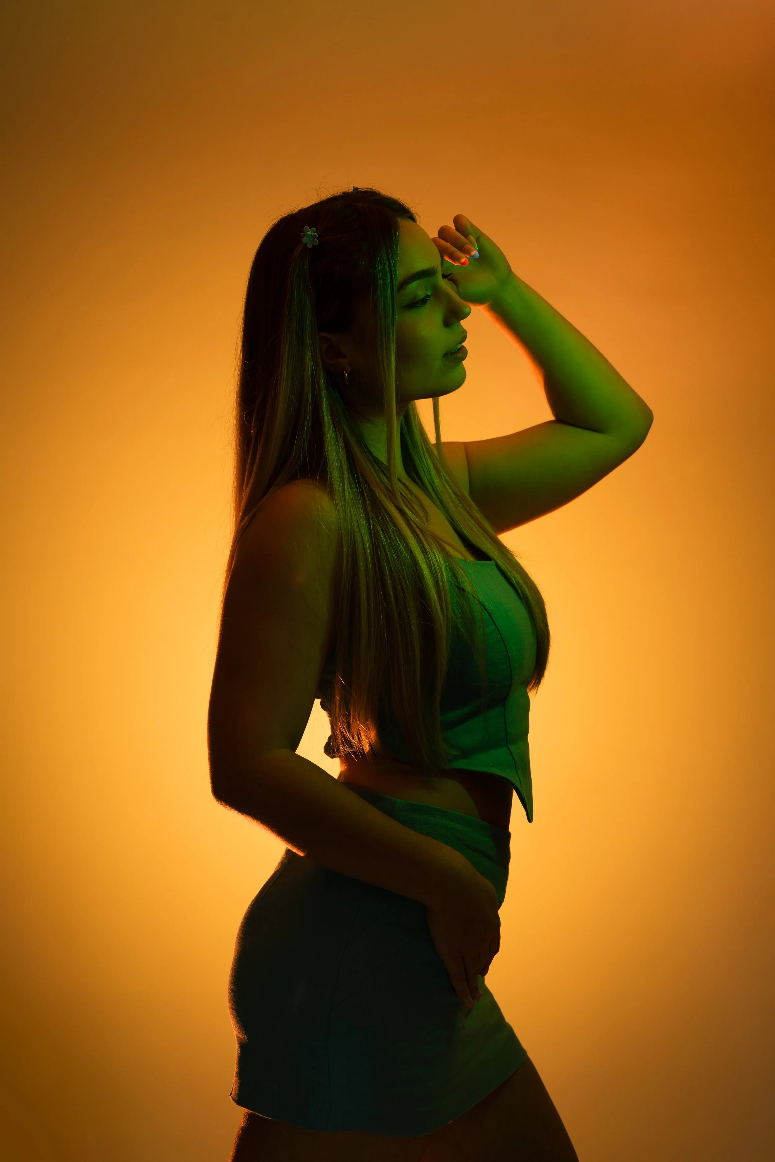 Silhouette of a woman with long hair, wearing a green top and skirt, posing with one arm raised and hand on her forehead, against a gradient yellow-orange background.