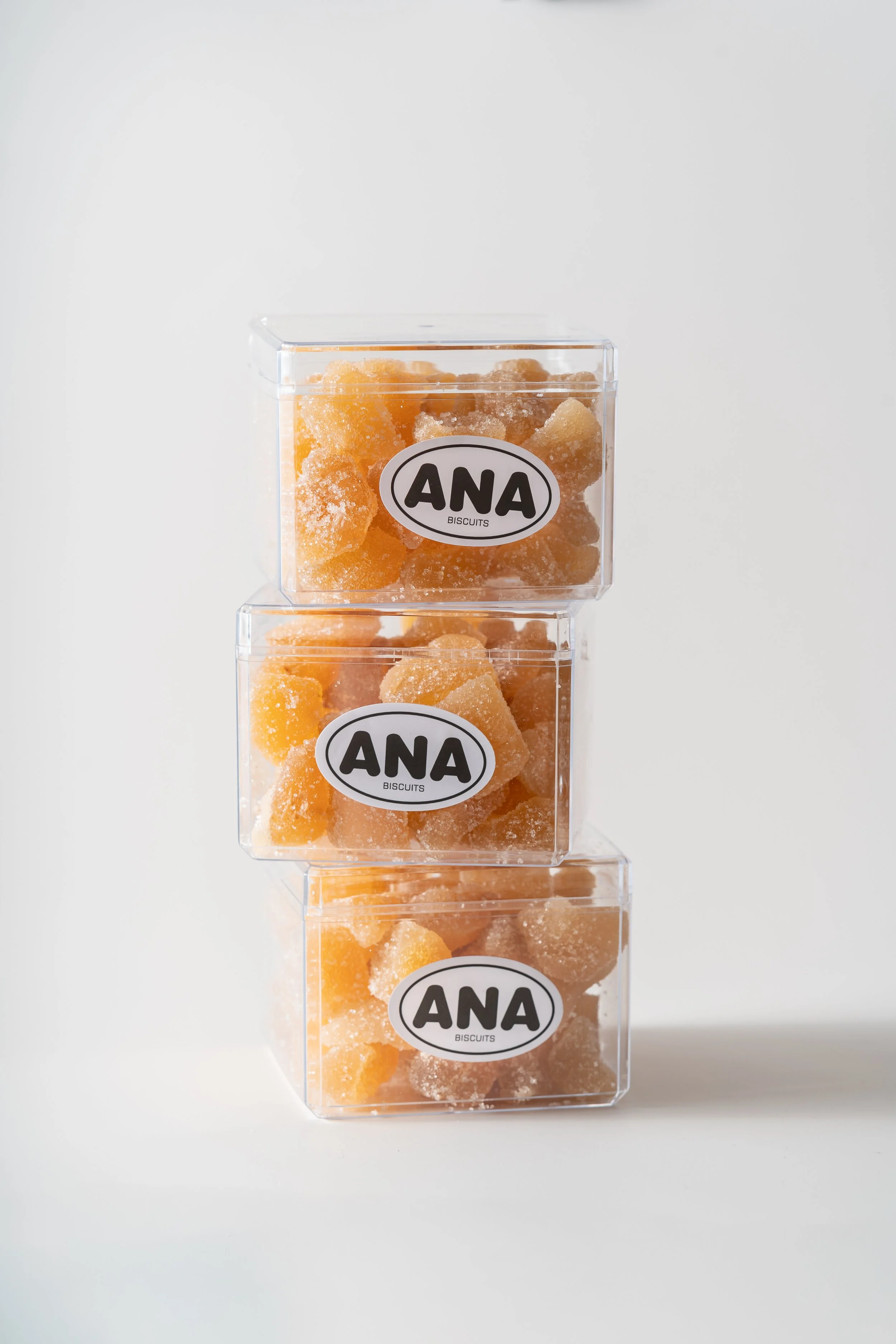 Three stacked clear plastic containers filled with orange-colored sugar-coated biscuits labeled 'ANA BISCUITS' against a white background.