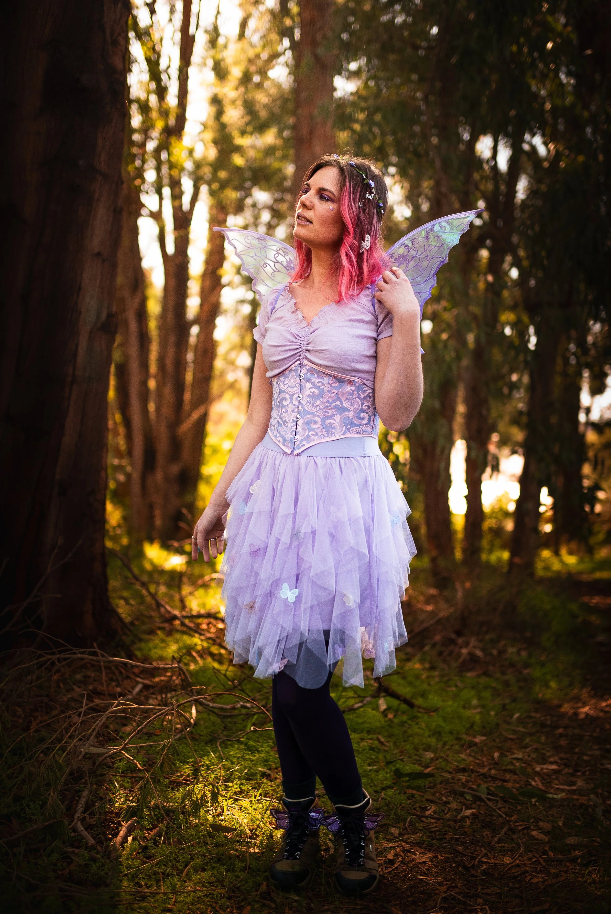 A woman dressed as a fairy in a purple, pink, and lavender costume with butterfly wings and a tutu skirt, standing in a forest with tall trees and dappled sunlight.