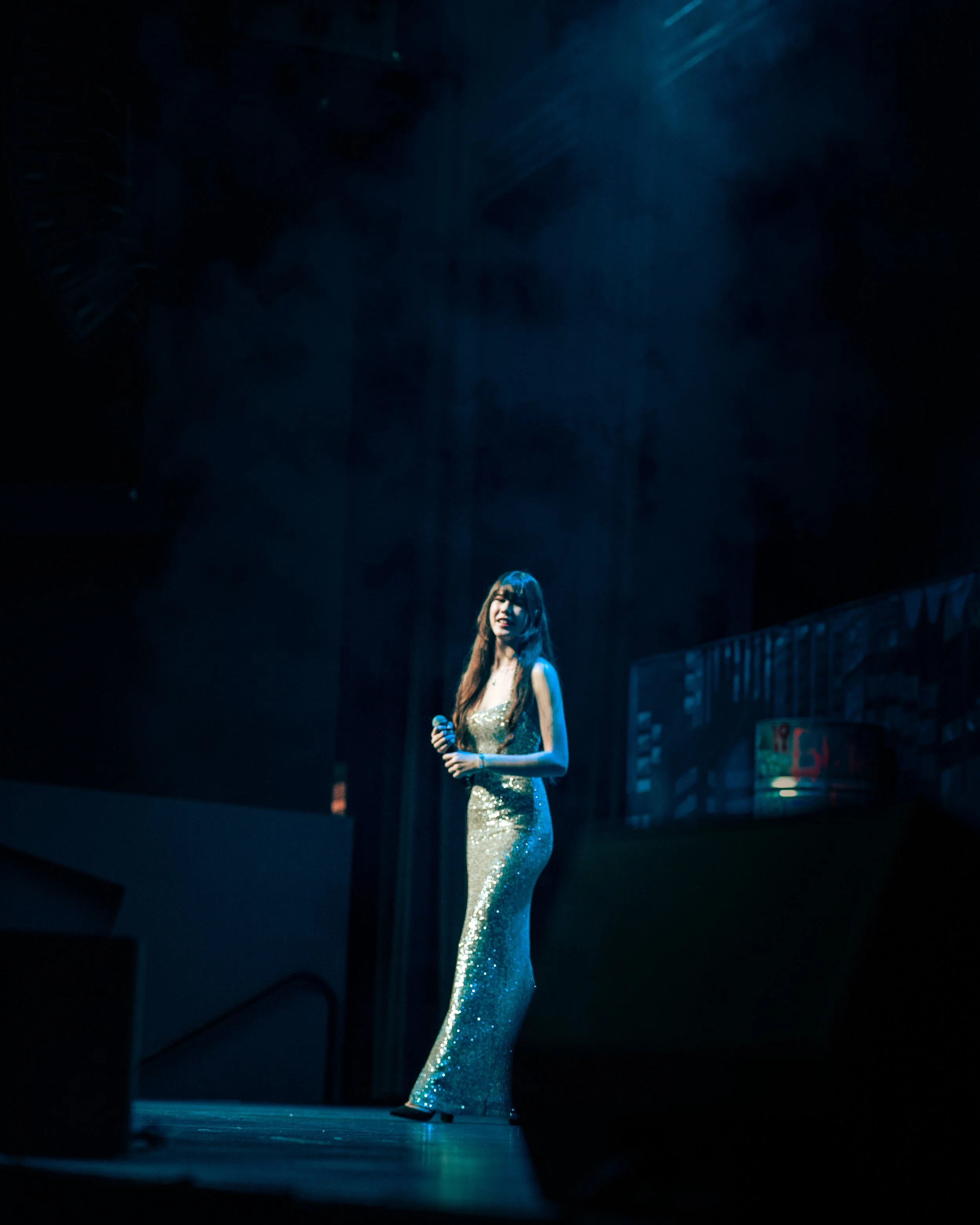 A woman wearing a shimmering gold gown holding a microphone stands on stage under blue lighting.