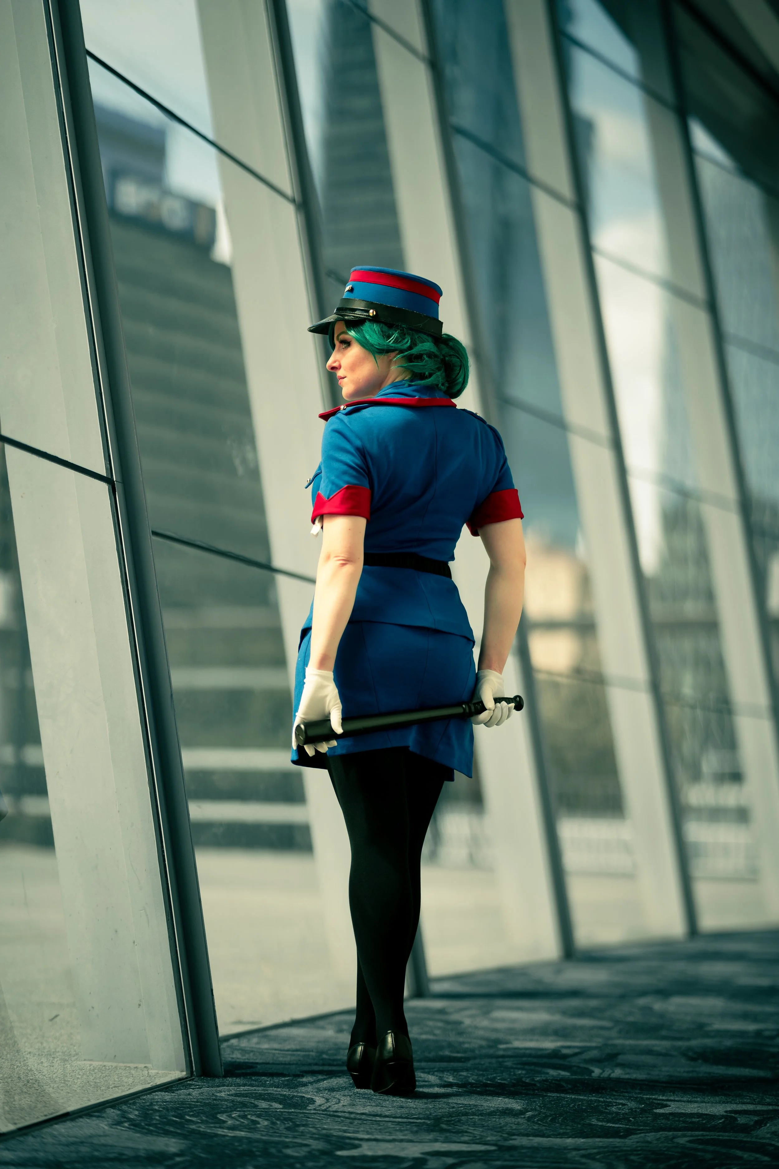 A woman dressed as a flight attendant with green hair, wearing a blue uniform with red accents, a hat, and holding a baton, standing outside a modern glass building.