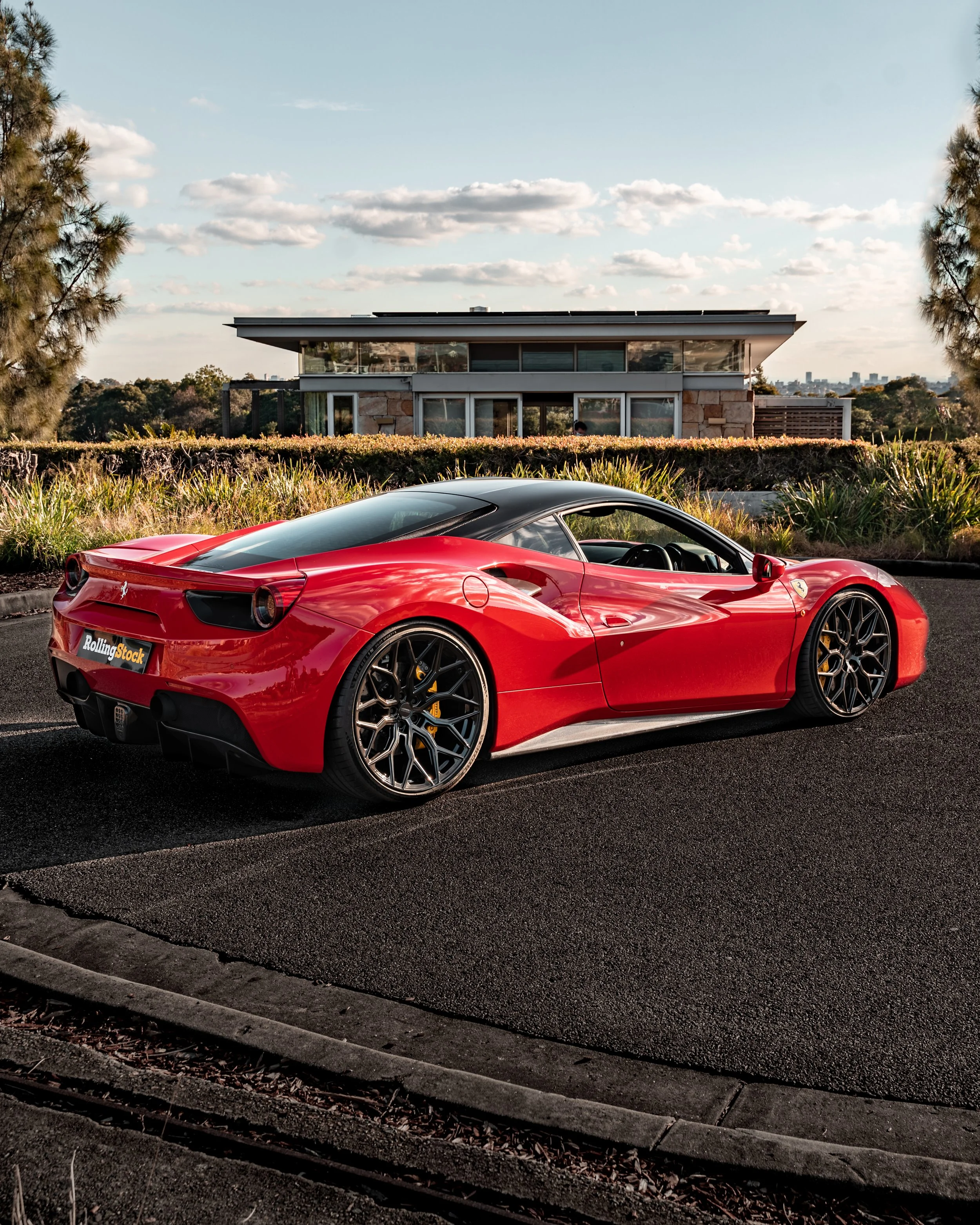 Red Ferrari sports car parked on a residential street with a modern house and trees in the background.