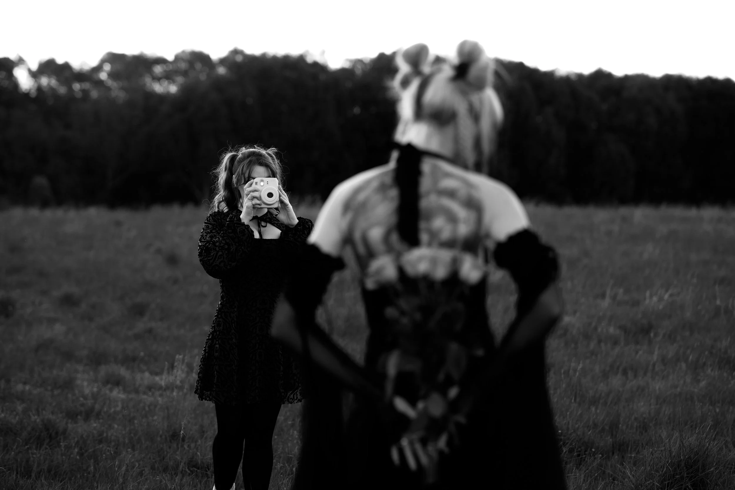 A girl in a dark dress takes a photograph of a person in a costume resembling a mythological or fantasy creature in a field with a hedge and trees in the background.