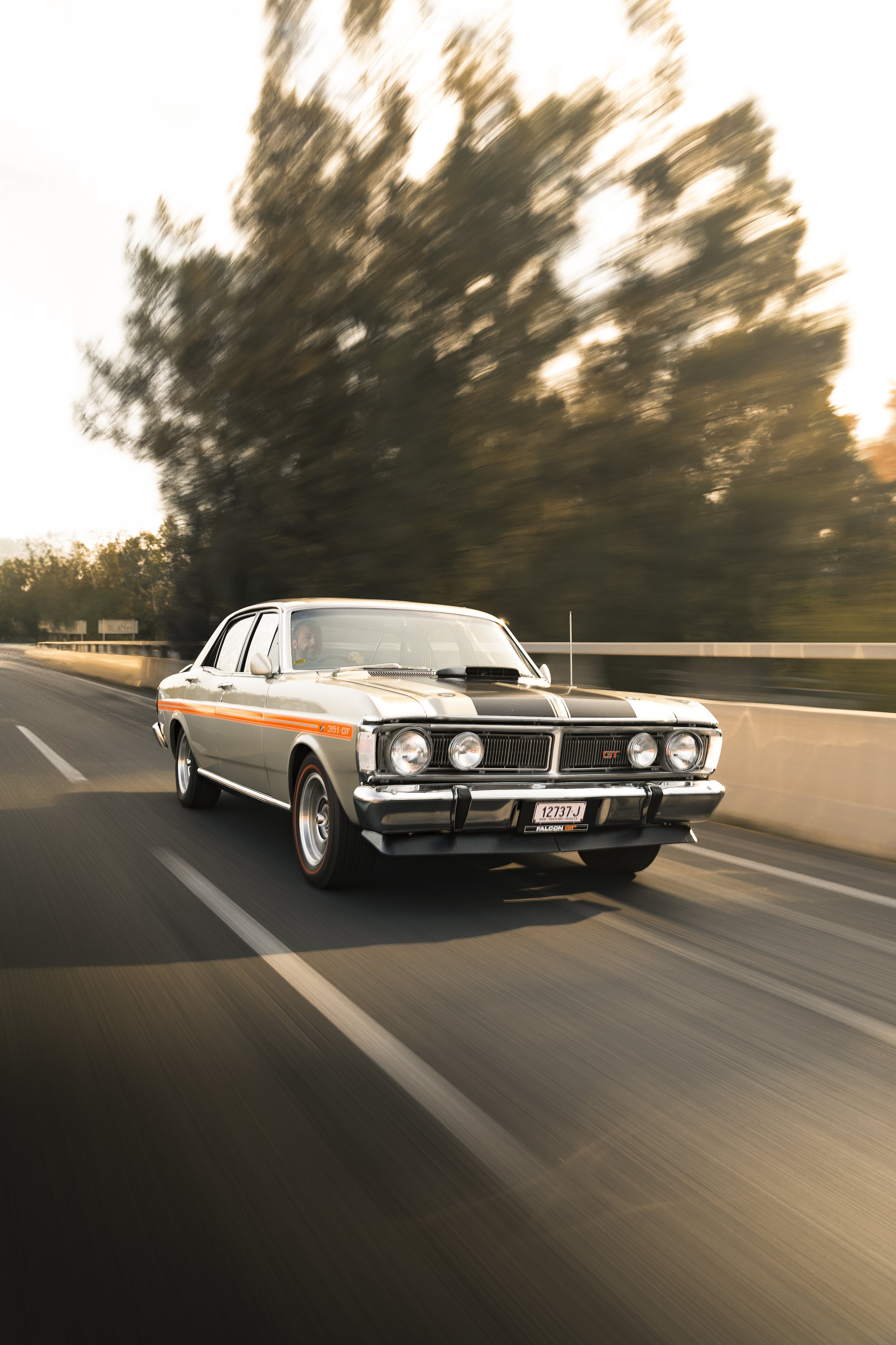 A vintage silver and black muscle car driving on a highway at sunset, with a blurred background of trees.
