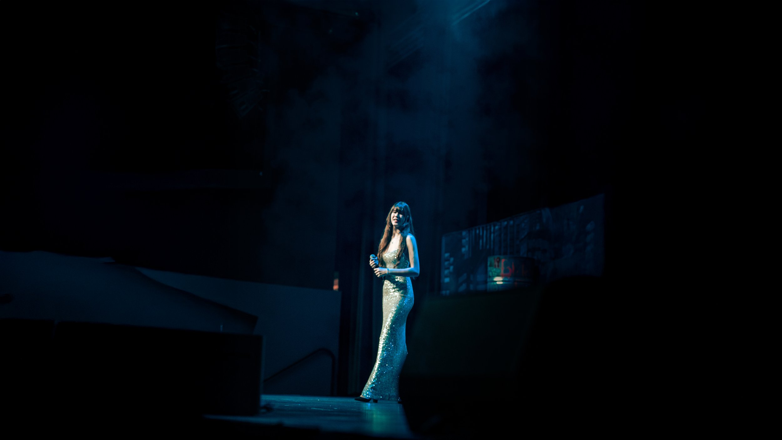 A woman in a long, shimmering gold gown holding a microphone, standing alone on a dimly lit stage with blue lighting.