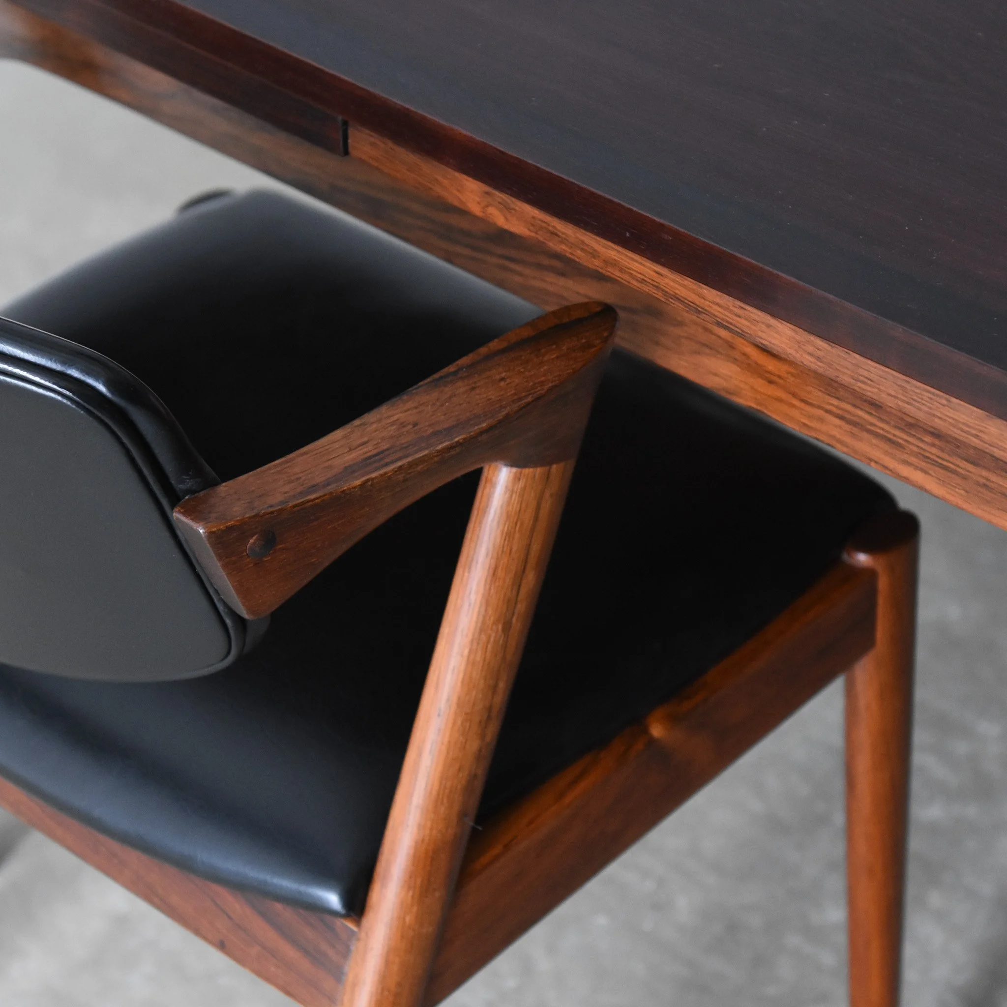 Close-up of a wooden chair with a black leather seat and armrest, placed under a wooden table.