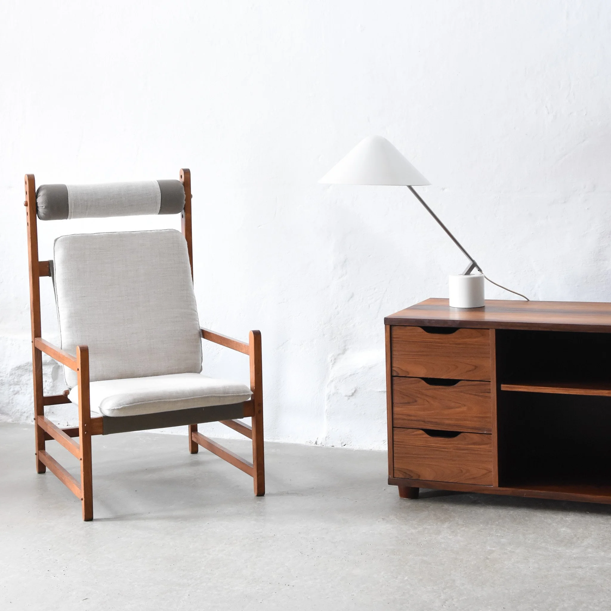 Minimalist room with a high-back wooden chair with gray cushion and headrest, and a wooden sideboard with a white table lamp on top, set against a white wall and concrete floor.