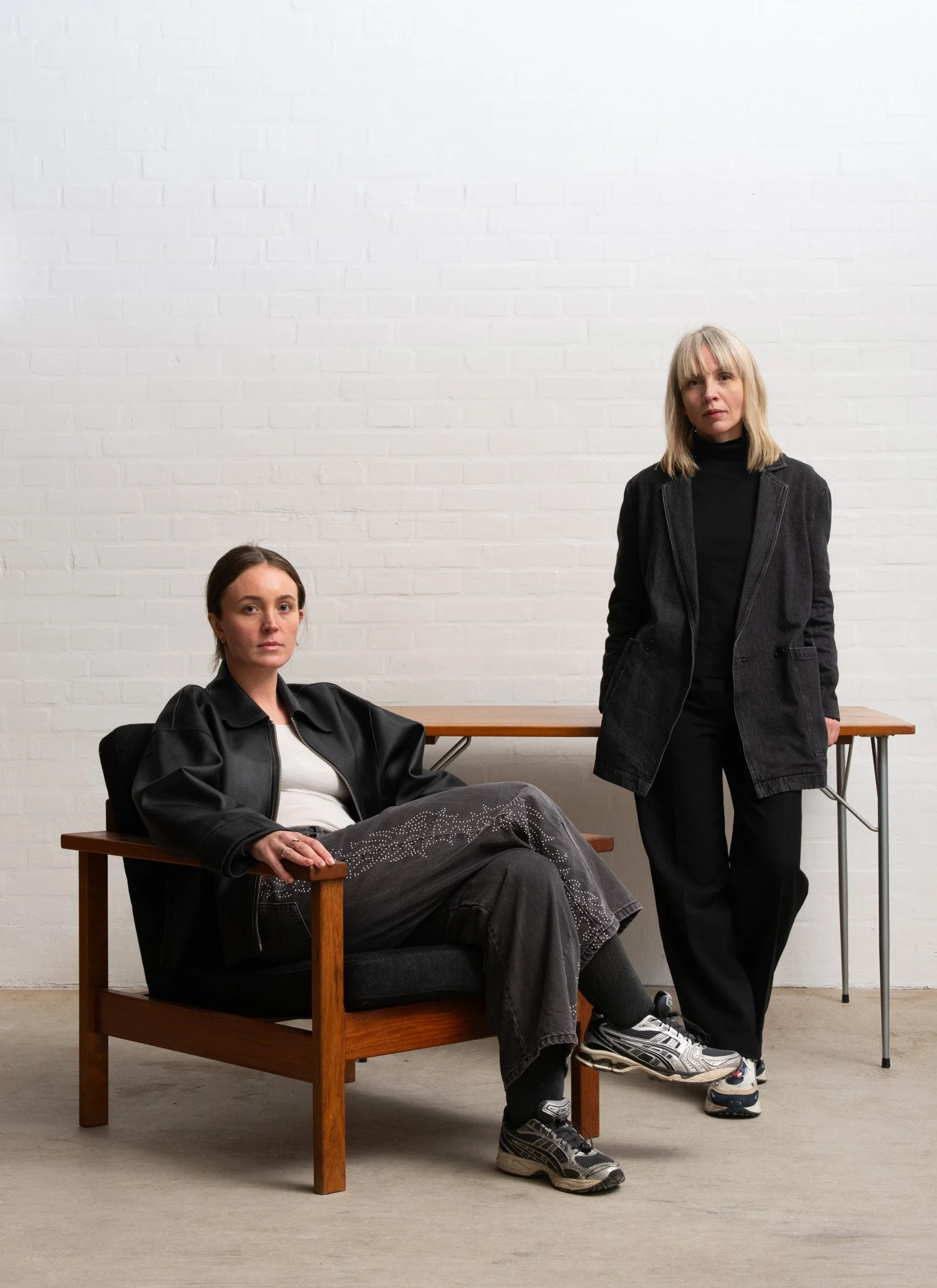 Two women in black clothing pose against a white brick wall, with one seated on a wooden chair and the other standing beside a wooden table.