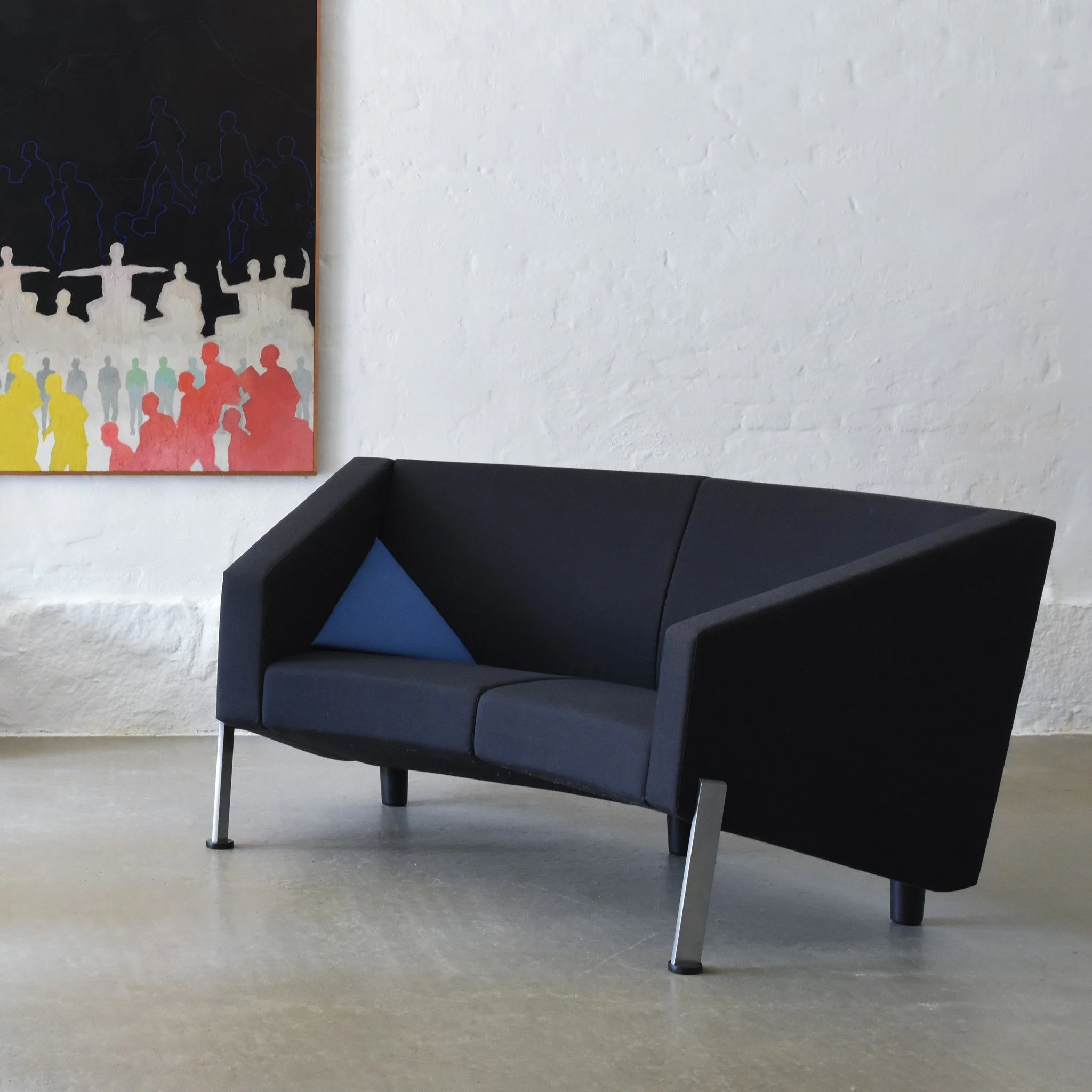 A modern black sofa with a blue pillow, placed in front of a white brick wall with a colorful abstract painting hanging on it.