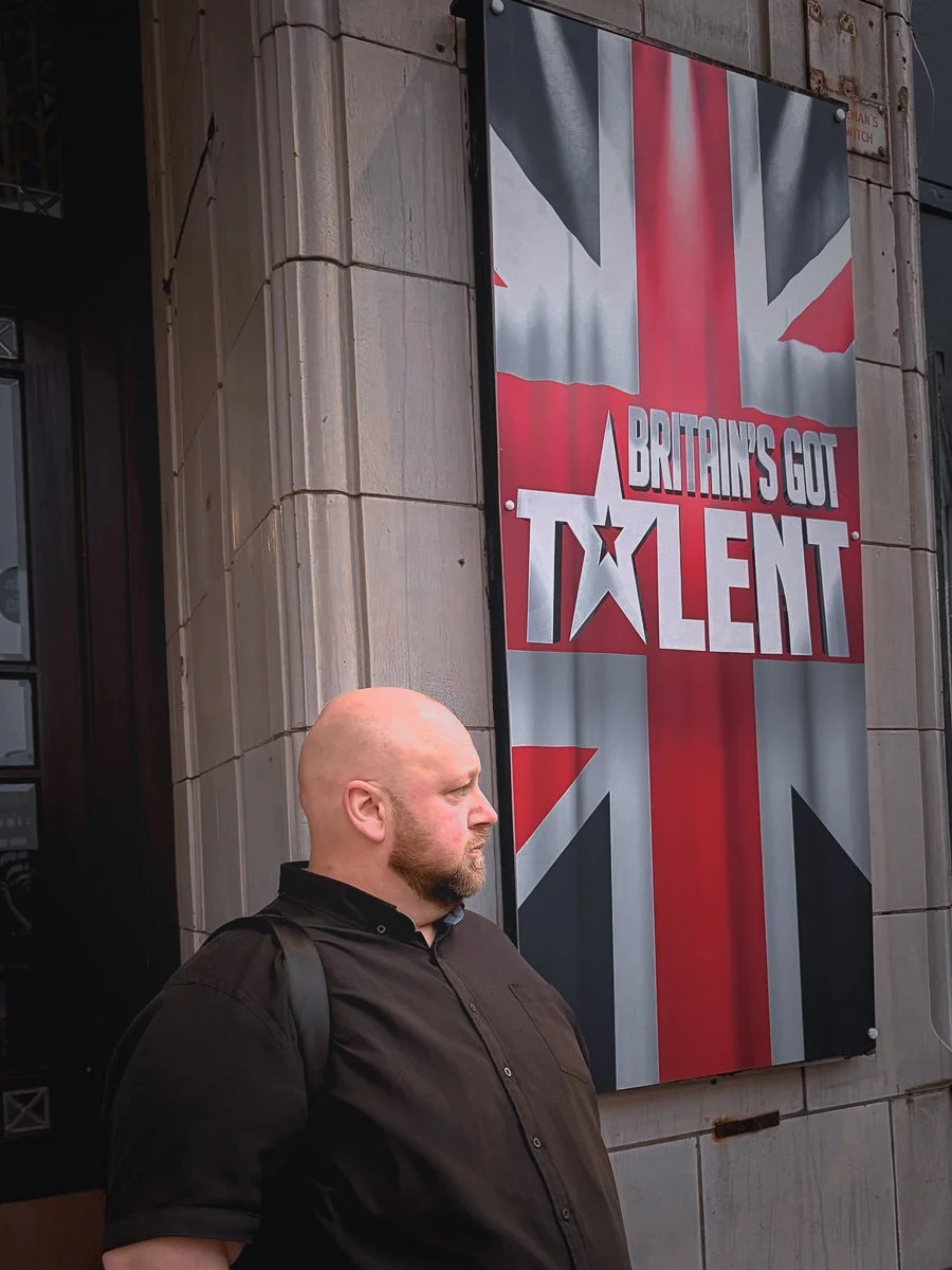 A man with a shaved head and beard standing outside a building next to a large sign that reads "Britain's Got Talent" with a Union Jack flag background.