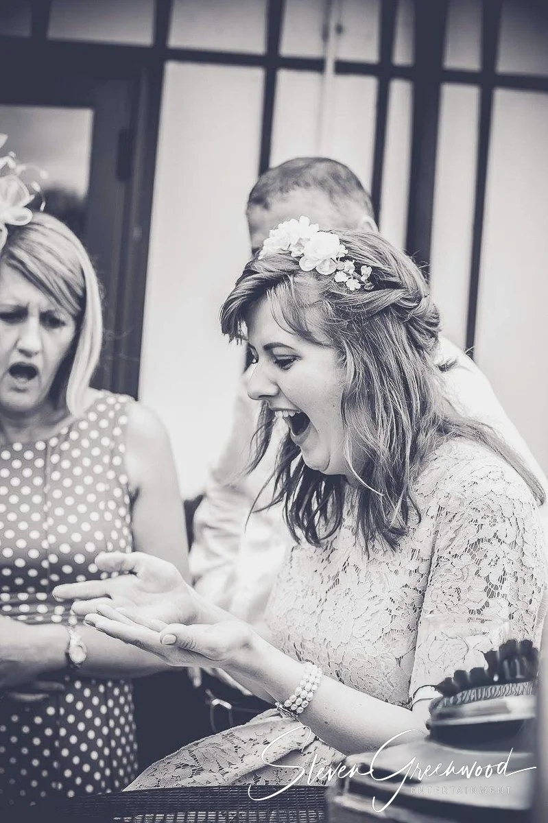 Two women and a man are celebrating, with the women appearing surprised and happy. One woman is wearing a polka dot dress and a floral headpiece, and the other woman is in a lace dress and also has a floral headpiece. Magician Steven Greenwood