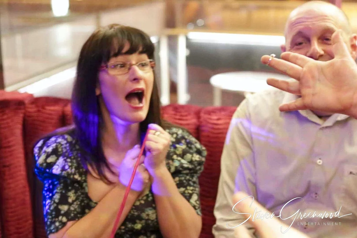 A woman with black hair and glasses is sitting on a red cushioned seat, appearing surprised or excited with her mouth open and hands clasped. A man with a shaved head is sitting next to her, Magician Steven Greenwood