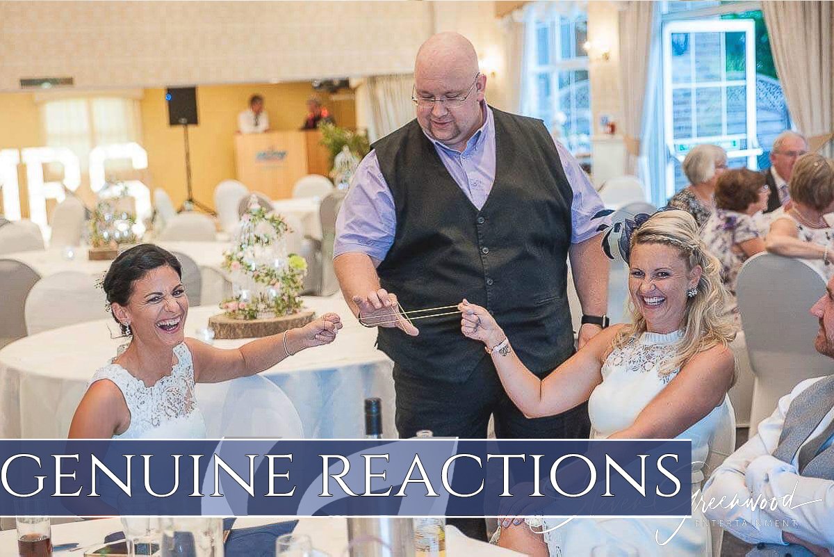 Happy women at a wedding reception, with one woman pulling a gag gift on a man, both women laughing, and a man watching. The scene is set in a decorated banquet hall with round tables and floral centerpieces, in front of a window with curtains.