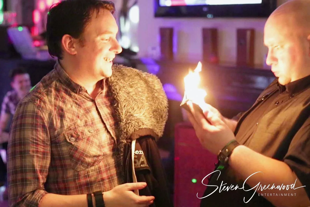 Two men in a lively bar or nightclub, one holding a lit match or small flame and the other smiling, with colorful lights in the background.