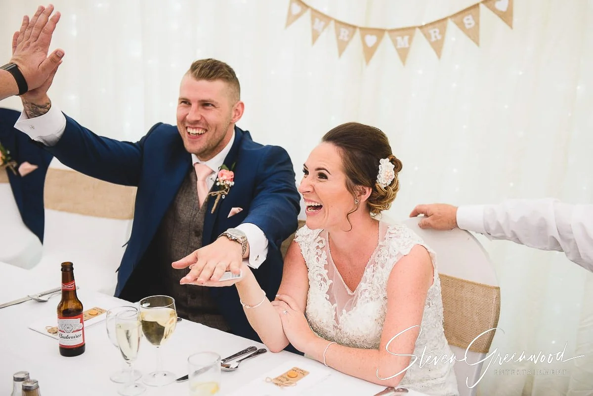 A wedding reception with a bride and groom sitting at a table, smiling and engaging with a guest. The bride is wearing a lace wedding dress and has a floral hair accessory. The groom is in a navy suit with a pink tie and boutonniere. There is a "MARR