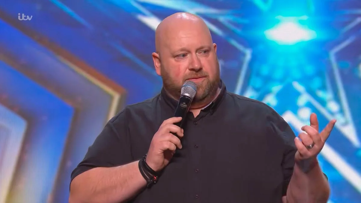 A man with a shaved head and beard holding a microphone on stage during a talent show.