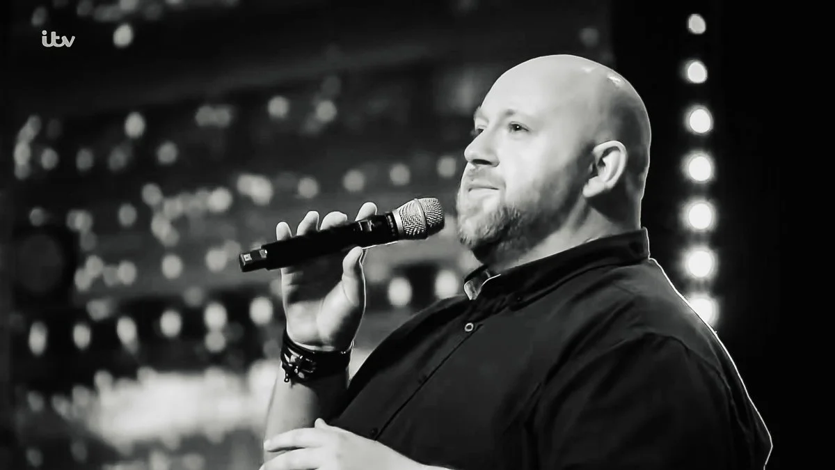 A man with a shaved head and beard is holding a microphone near his mouth, standing on a stage with bright lights in the background, speaking or singing.
