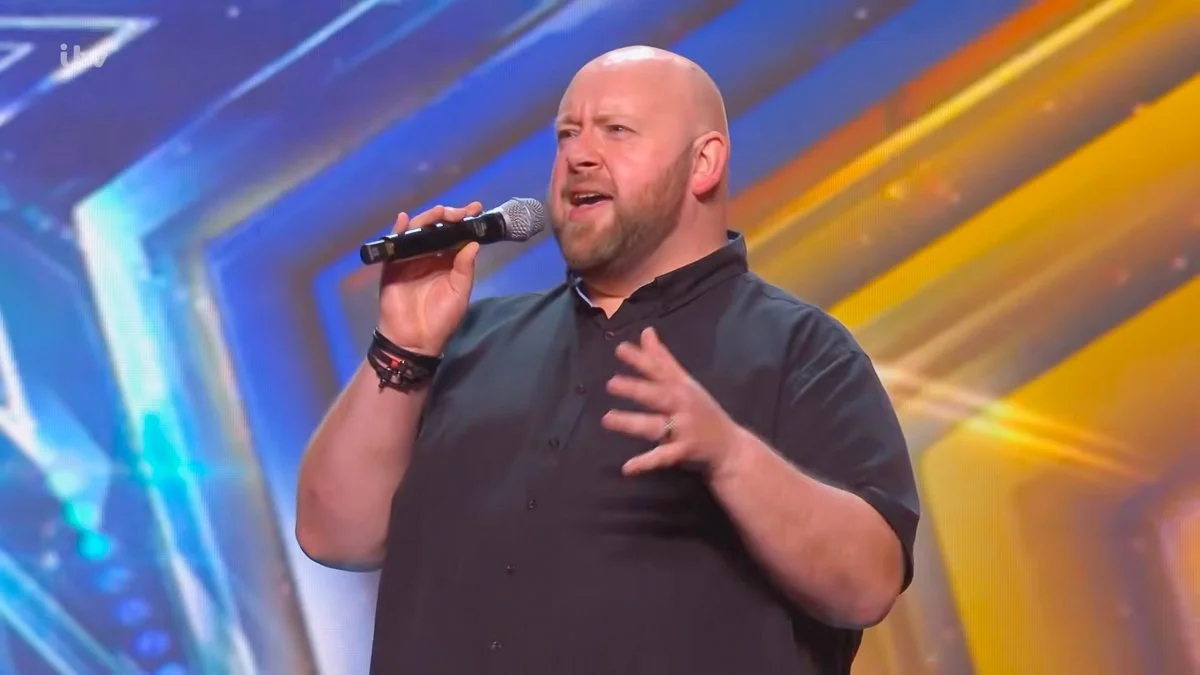 A man with a beard and bald head singing into a microphone on stage with colorful blue, yellow, and orange background lights.