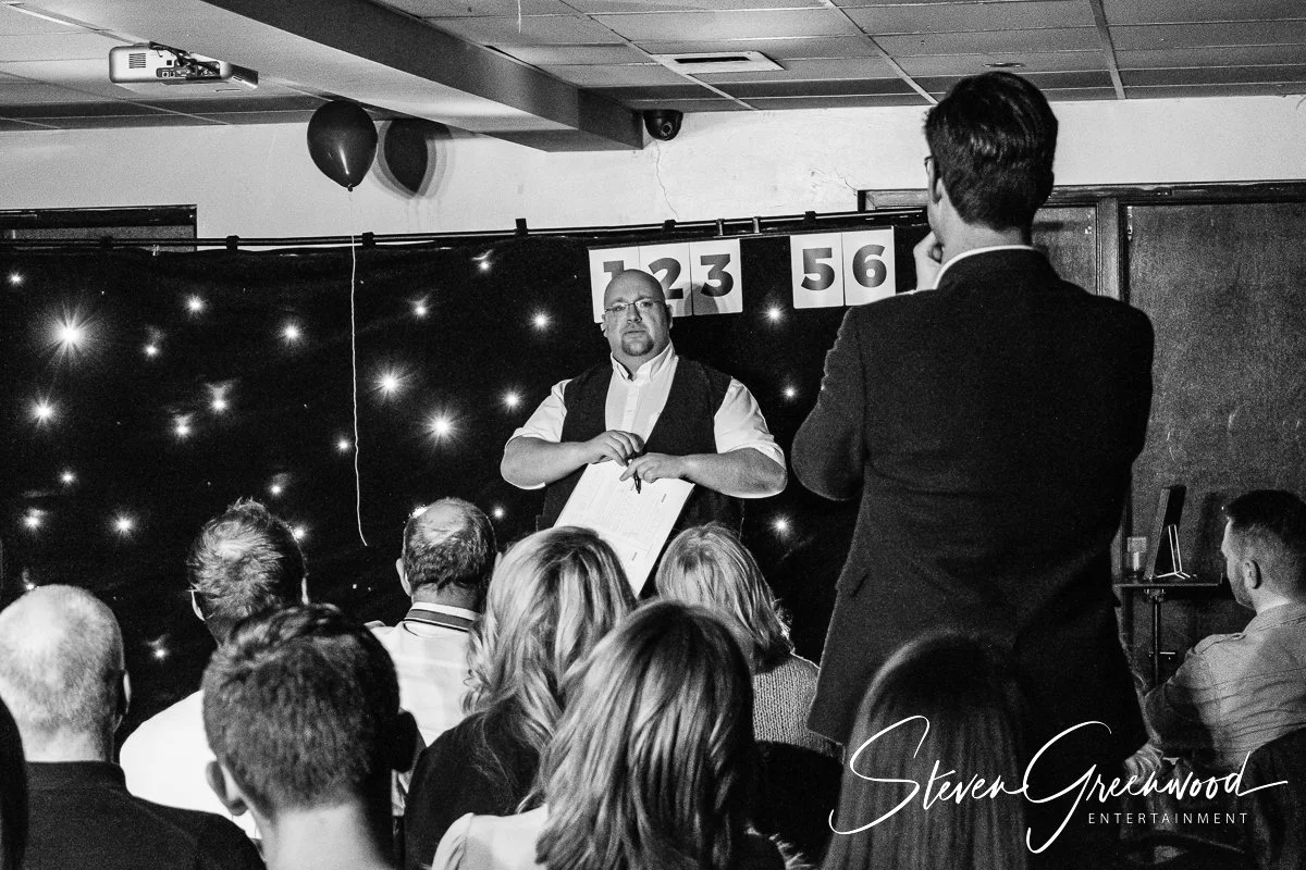 Man presenting to an audience at an event, with a backdrop decorated with star-like lights and numbered banners, and a person standing and facing the presenter. Magician Steven Greenwood