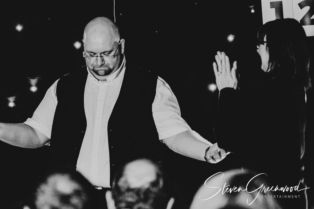 A man and woman dancing together, with the woman raising her hand in front of her face. The setting appears to be a nighttime event, with a dark background and some bright spots. The image is in black and white. Magician Steven Greenwood