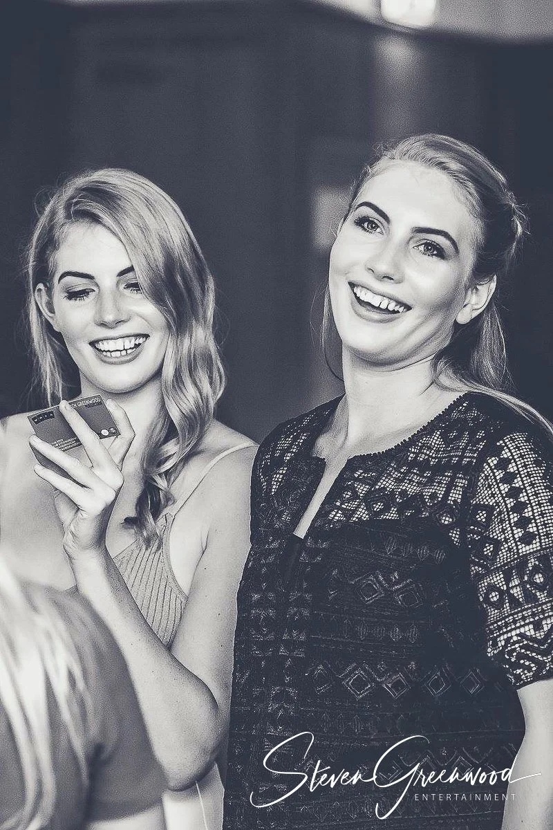 Two young women smiling and enjoying themselves. One woman is holding a smartphone. Magician Steven Greenwood