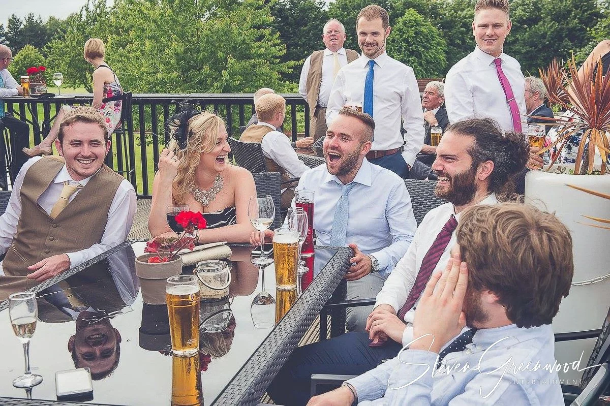 People in formal attire enjoying drinks and laughing at an outdoor social event on a balcony with greenery in the background. Magician Steven Greenwood
