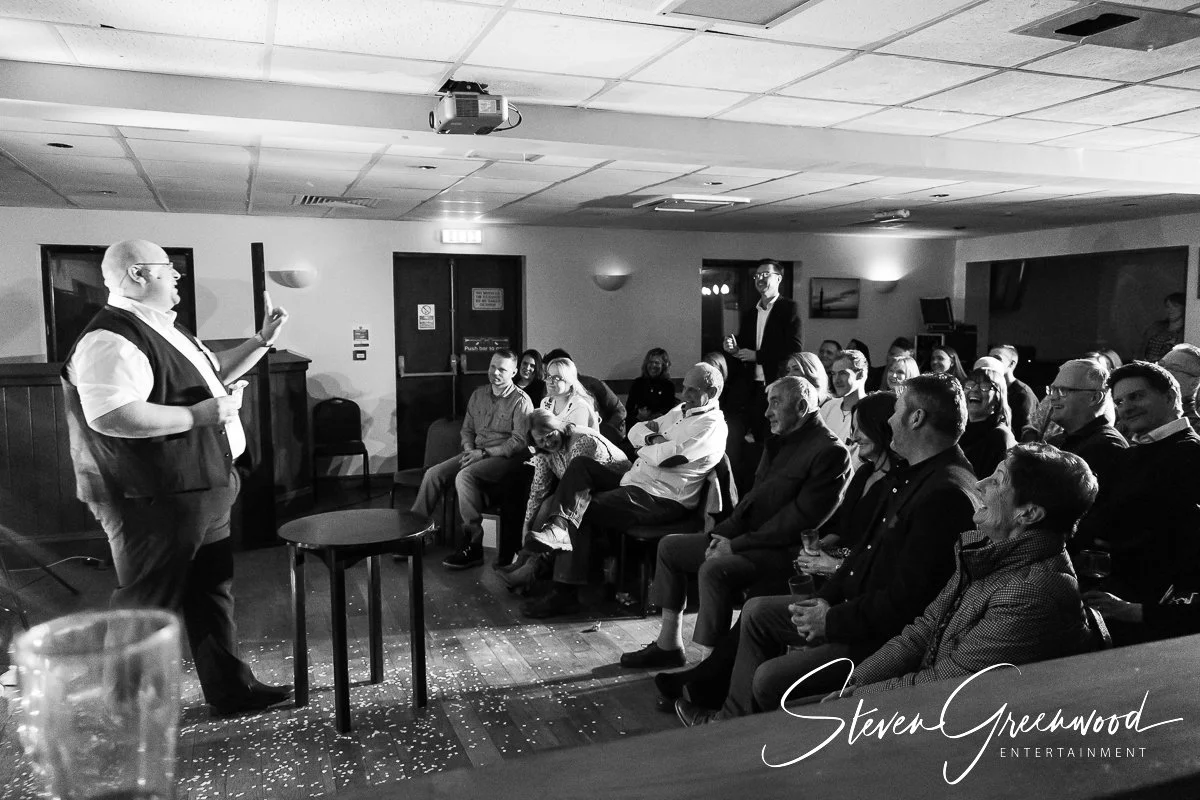 A man giving a speech in front of an audience at an entertainment event, with some audience members smiling and laughing, in a dimly lit room.