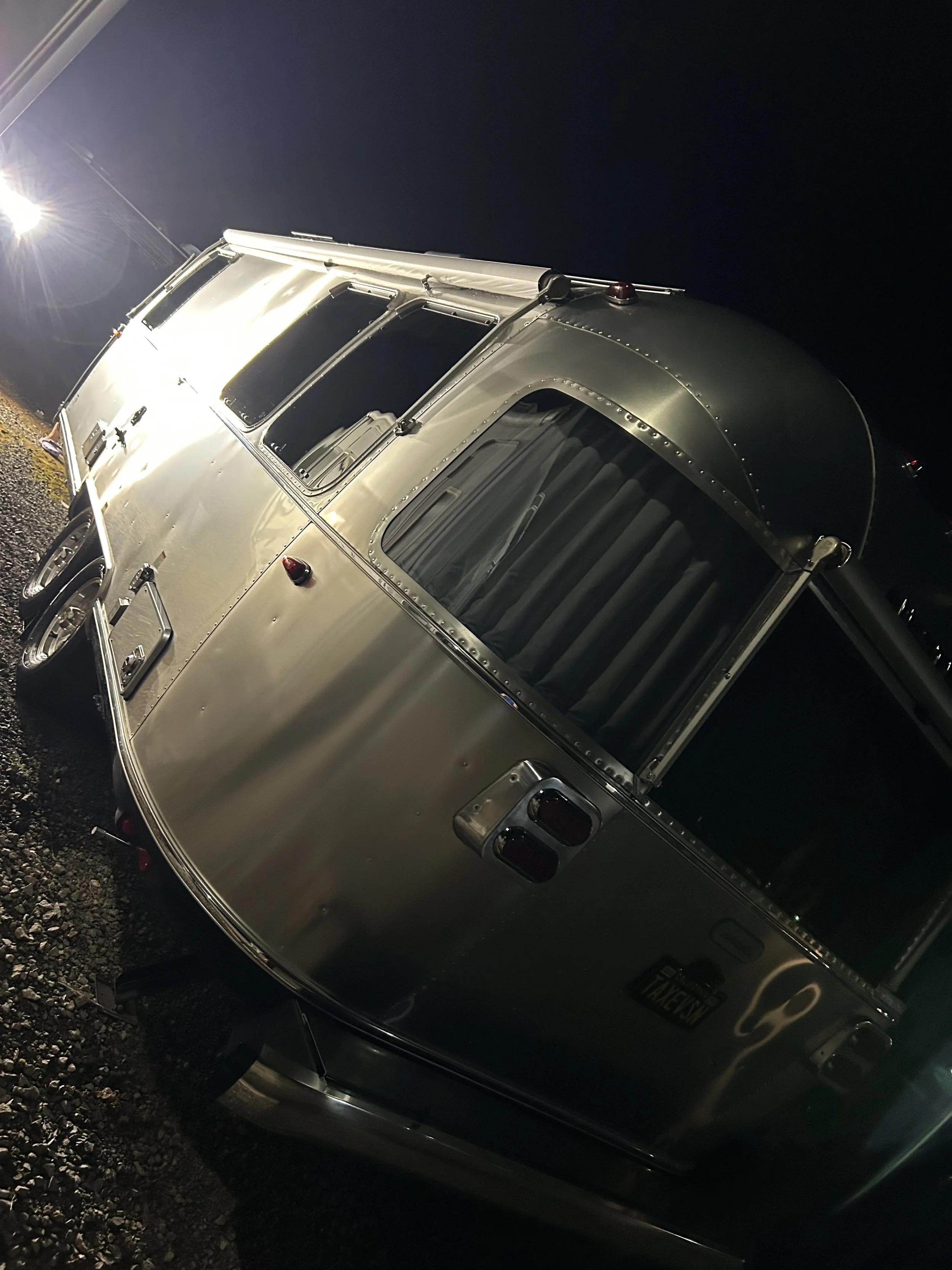 Modern wooden building with a large glass window, surrounded by yellowish-green plants, set against a backdrop of mountains with grassy and rocky terrain.
